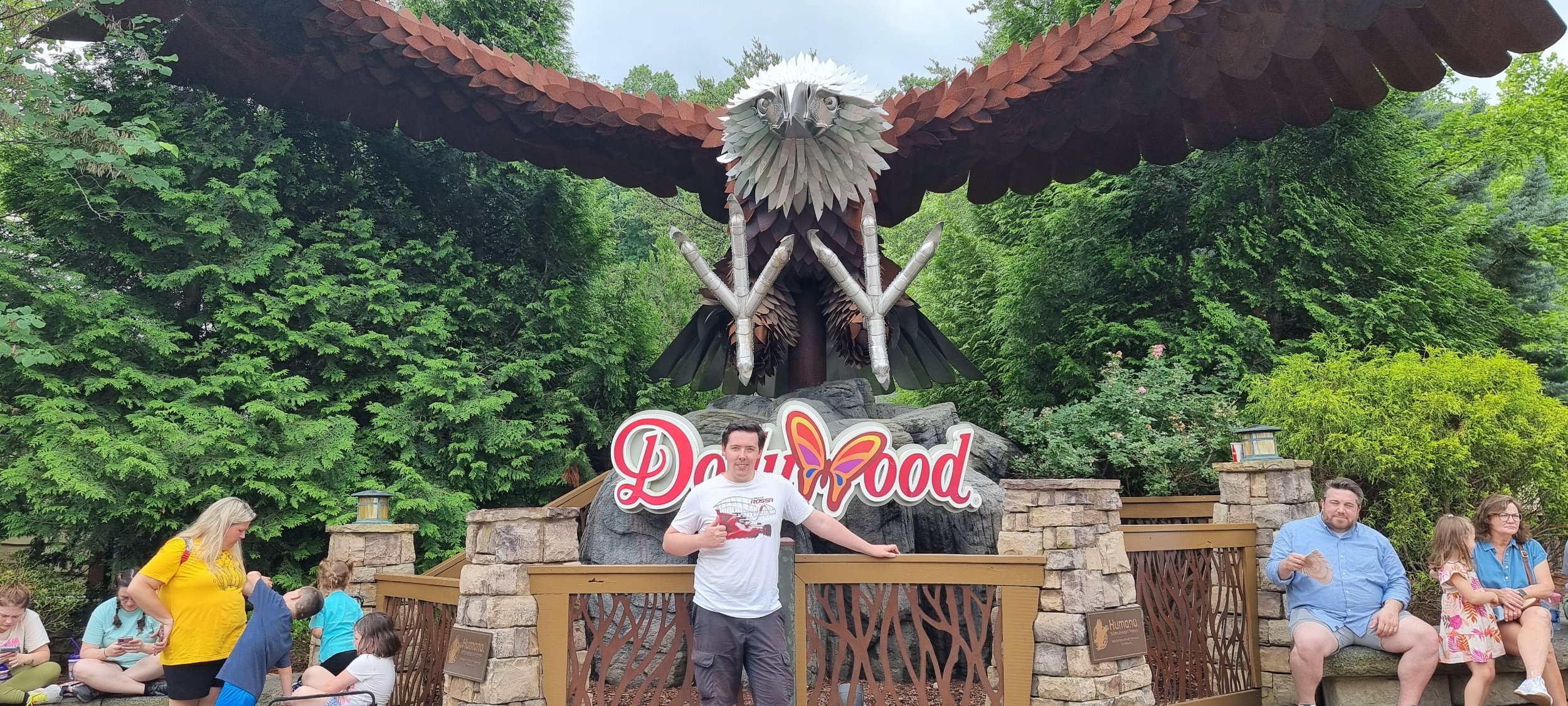 A family of five sitting on a bench and a man standing in front of a large metal sculpture of an eagle at Dollywood amusement park. The eagle has its wings spread wide and is perched on rocks. The background features green trees and bushes.