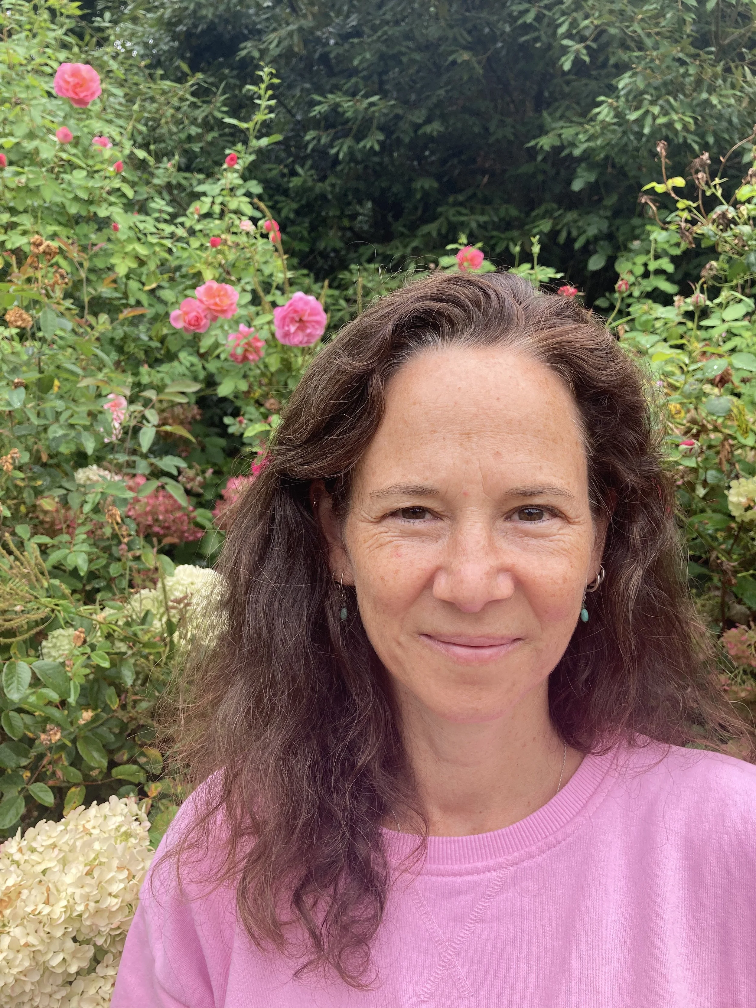 A woman with long, wavy brown hair wearing a pink sweatshirt and turquoise earrings stands in front of a bush with pink and white flowers.