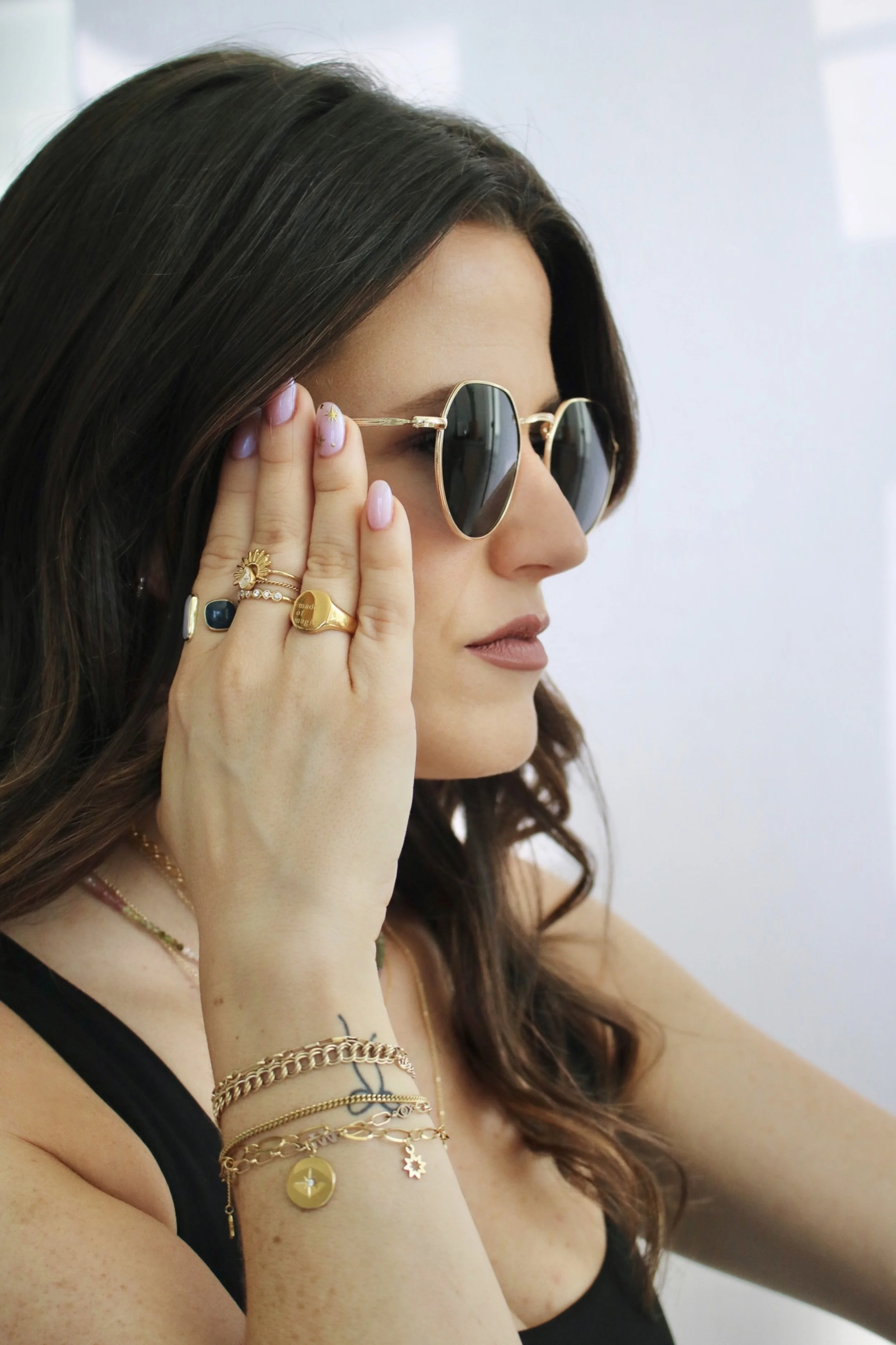 A woman with dark wavy hair wearing sunglasses, multiple rings, and layered gold bracelets, touching her face with her hand.