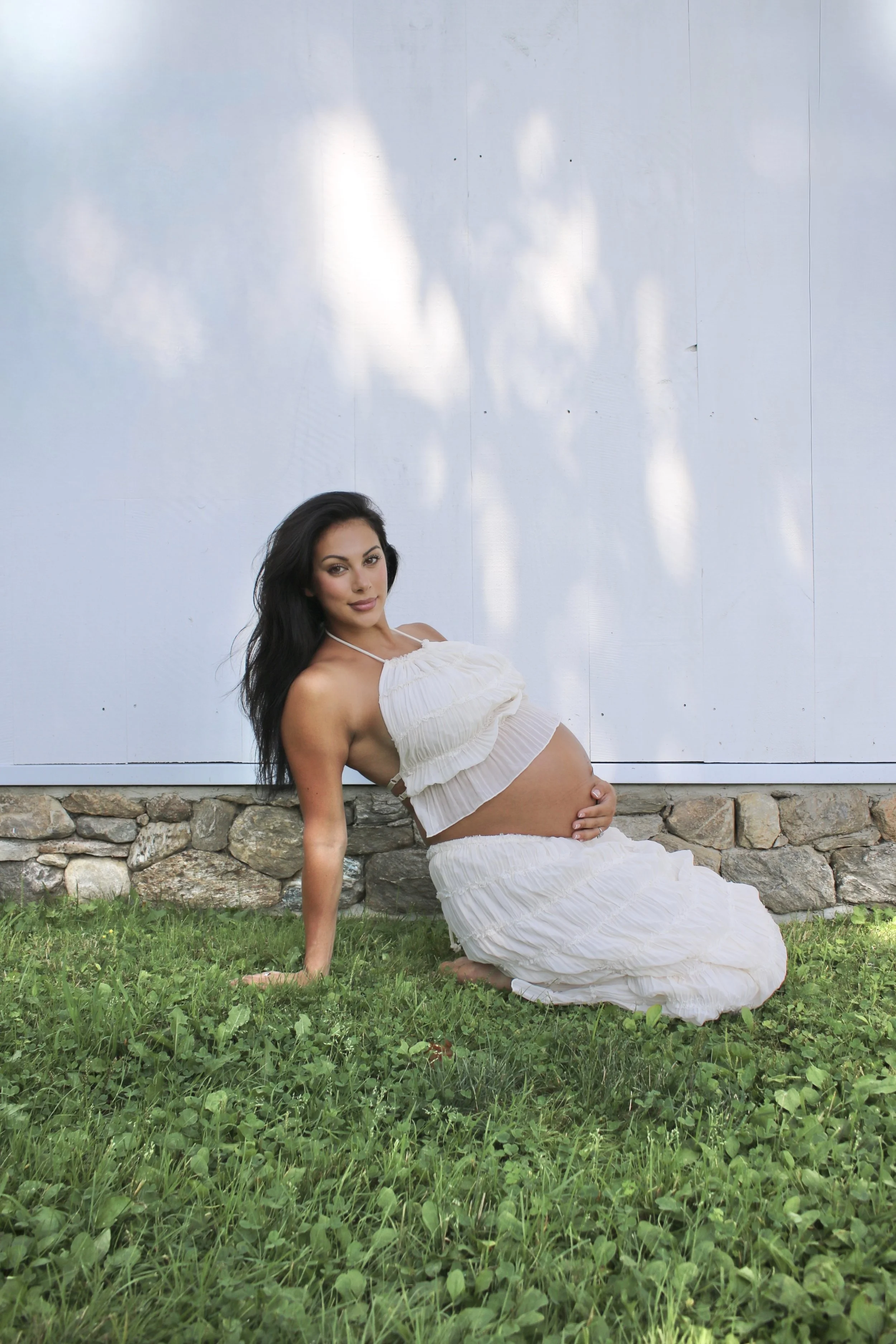 A pregnant woman with dark hair, wearing a white ruffled dress, sitting on grass with a stone border behind her, posing against a white wall with shadows of leaves.