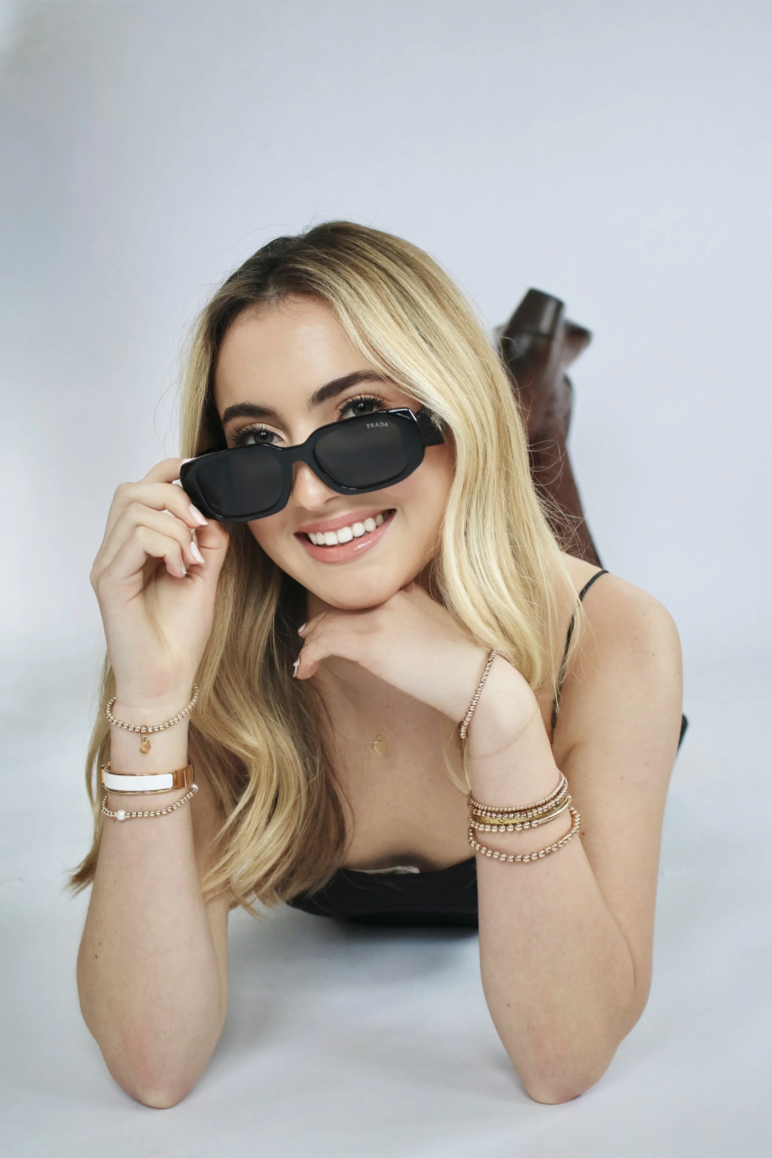 A woman with blonde hair, wearing sunglasses, smiling, lying on her stomach with her chin resting on her hand, wearing jewelry and a black top.