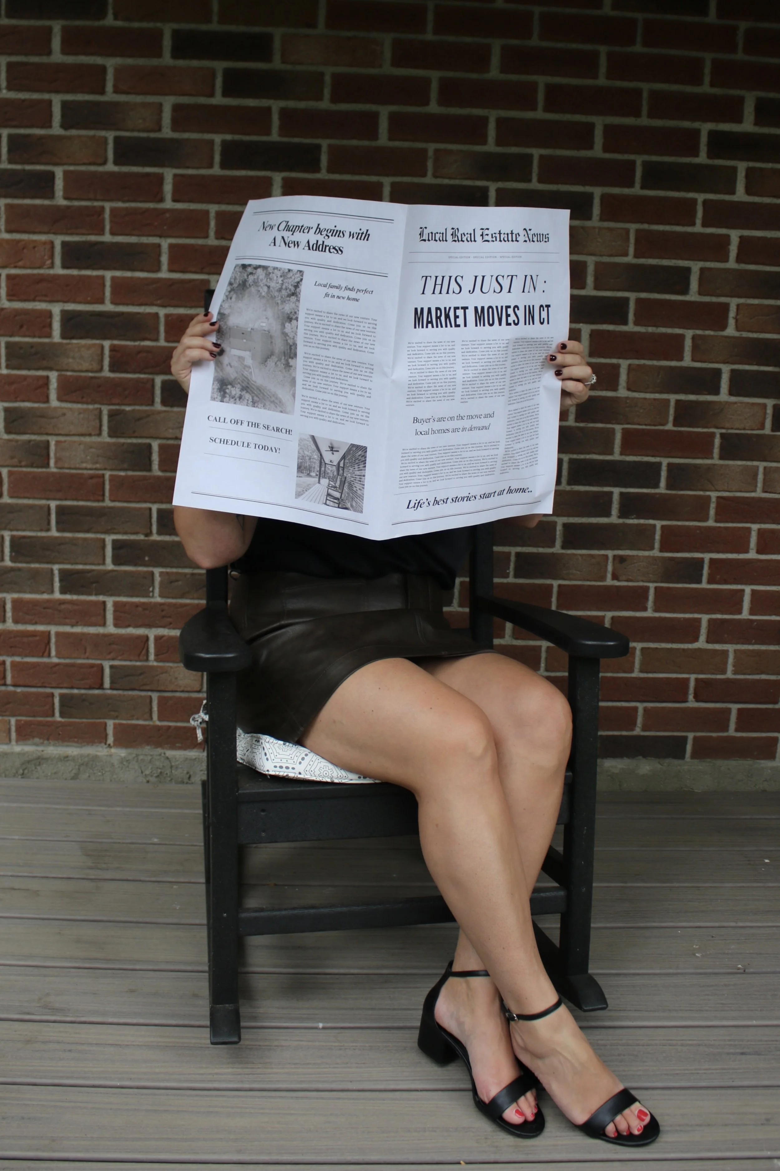 A woman sitting on a black chair outdoors against a brick wall, reading a newspaper covering her face.