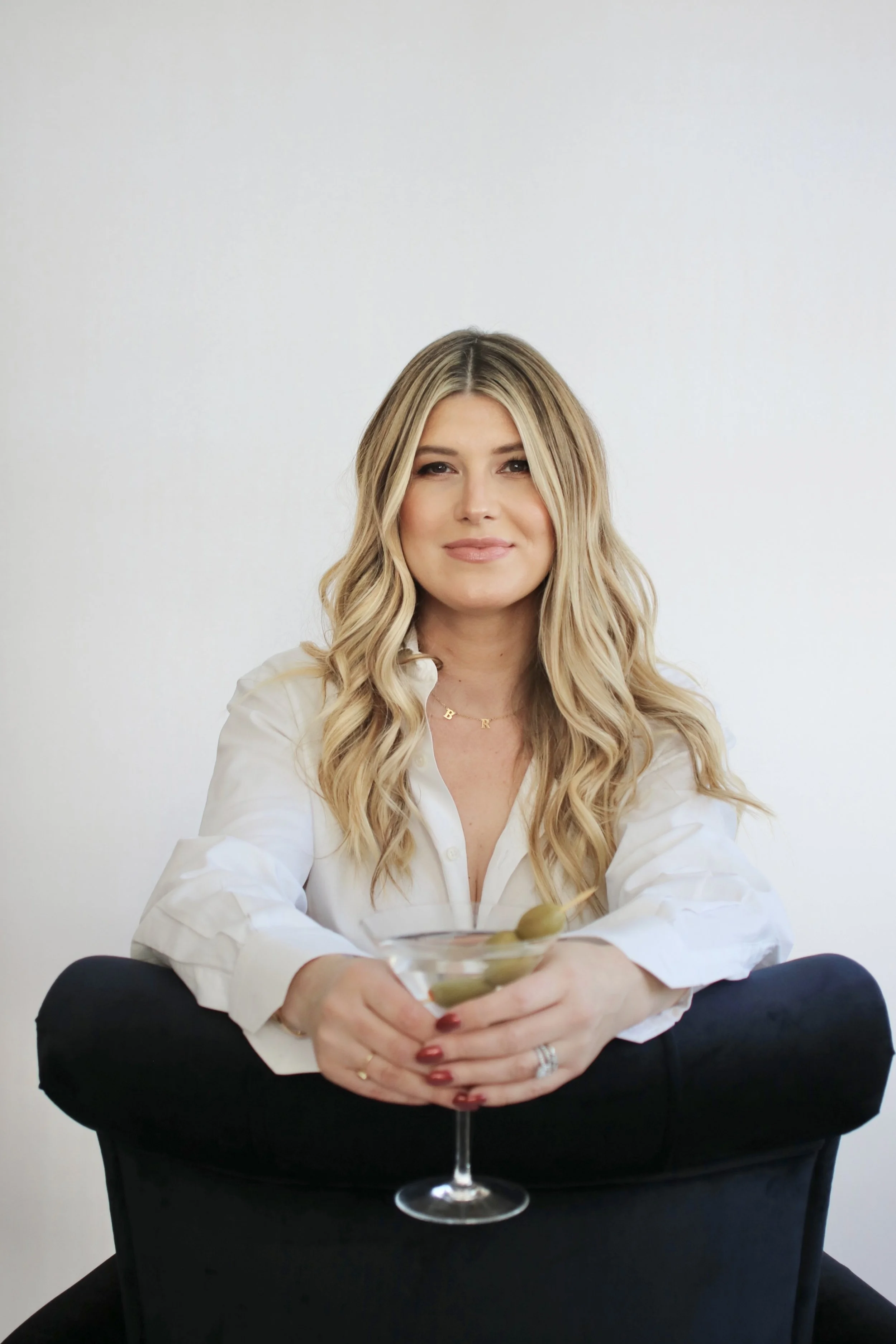 A woman with long blonde hair, wearing a white shirt, holding a glass of martini with olives, sitting in a black chair against a plain white background.