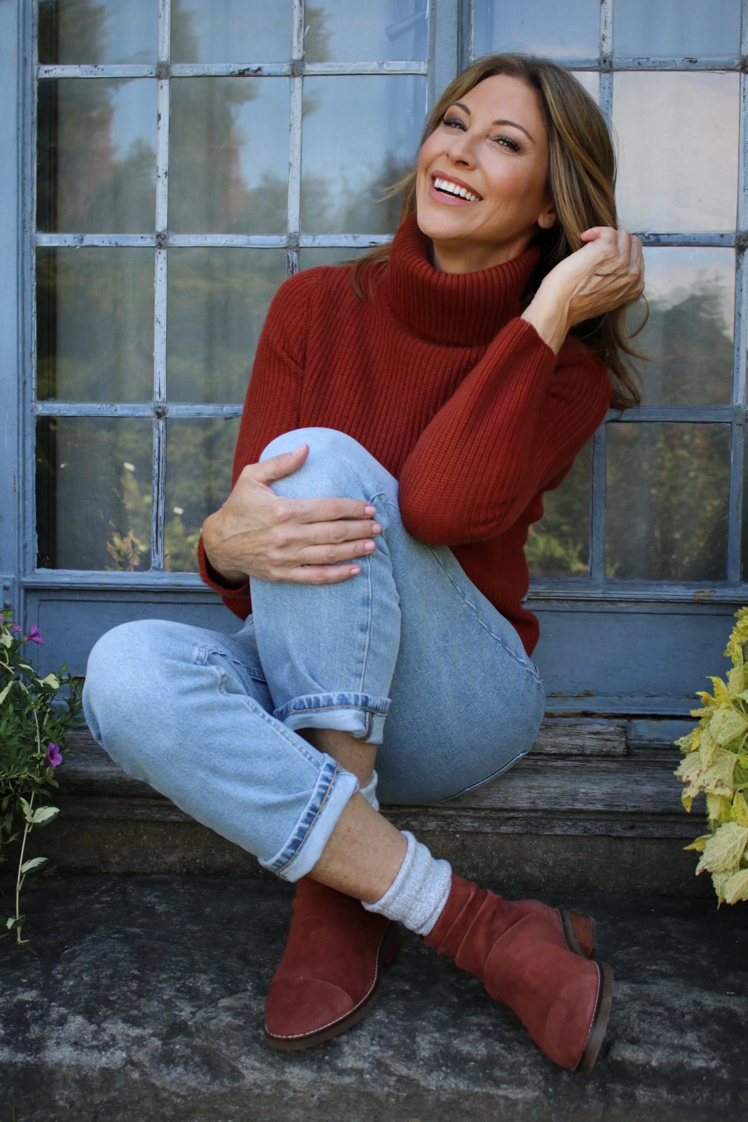 A woman sitting on a step in front of a blue window, wearing a red turtleneck sweater, light blue jeans, gray socks, and red boots, smiling and touching her hair.