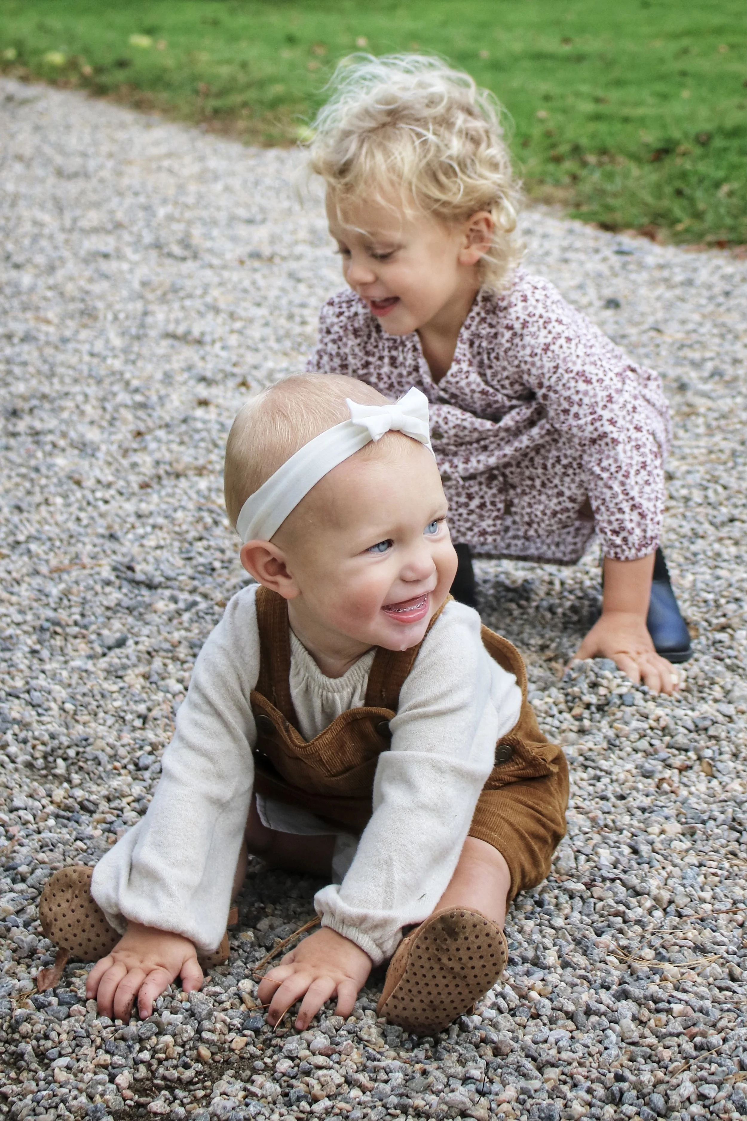 Two young children playing on gravel outdoors, one crawling with a headband and the other crouching behind, smiling and interacting.