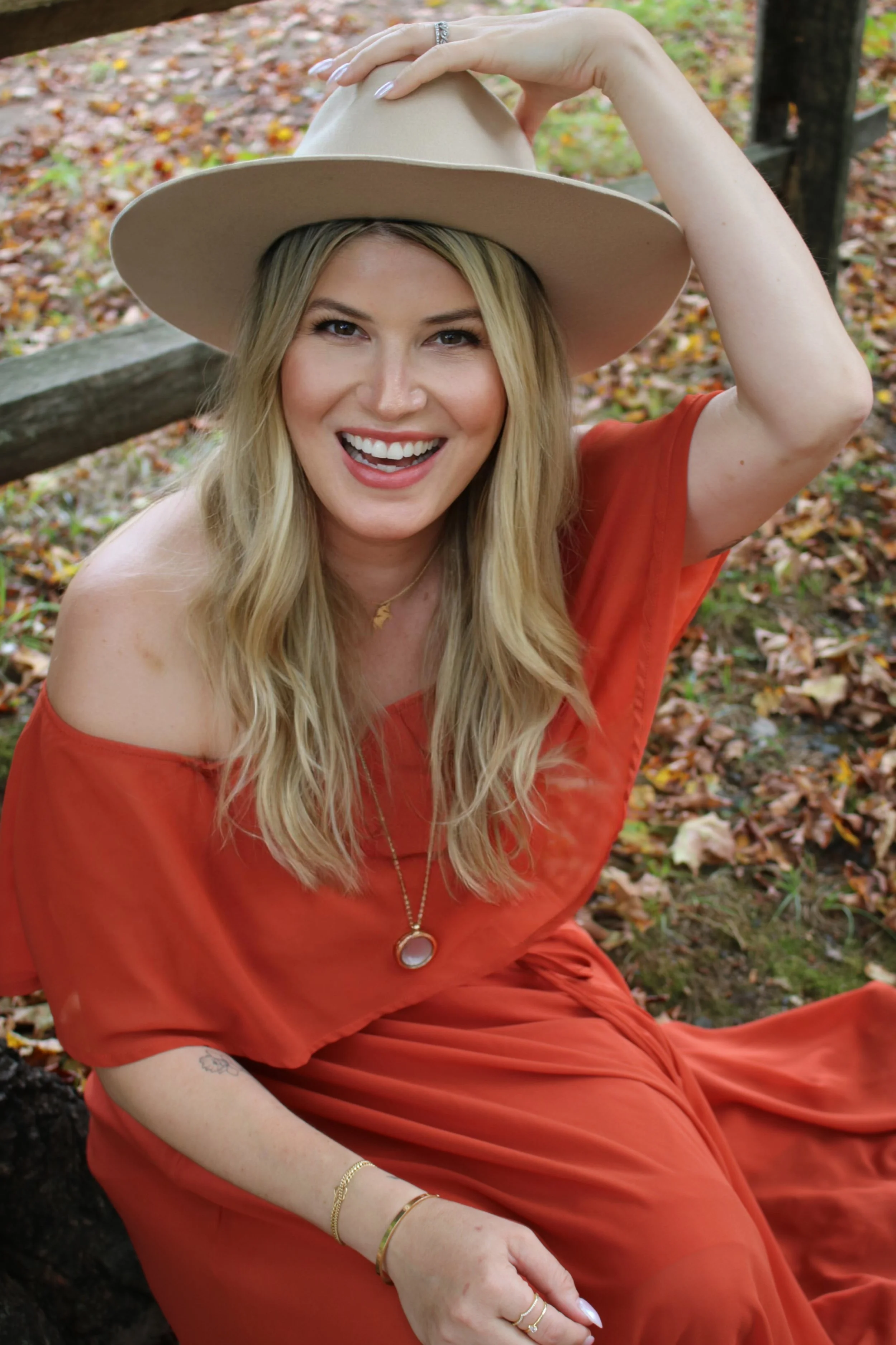 A smiling woman with long blonde hair, wearing an orange off-the-shoulder dress and a large beige hat, sitting outdoors on fallen autumn leaves.