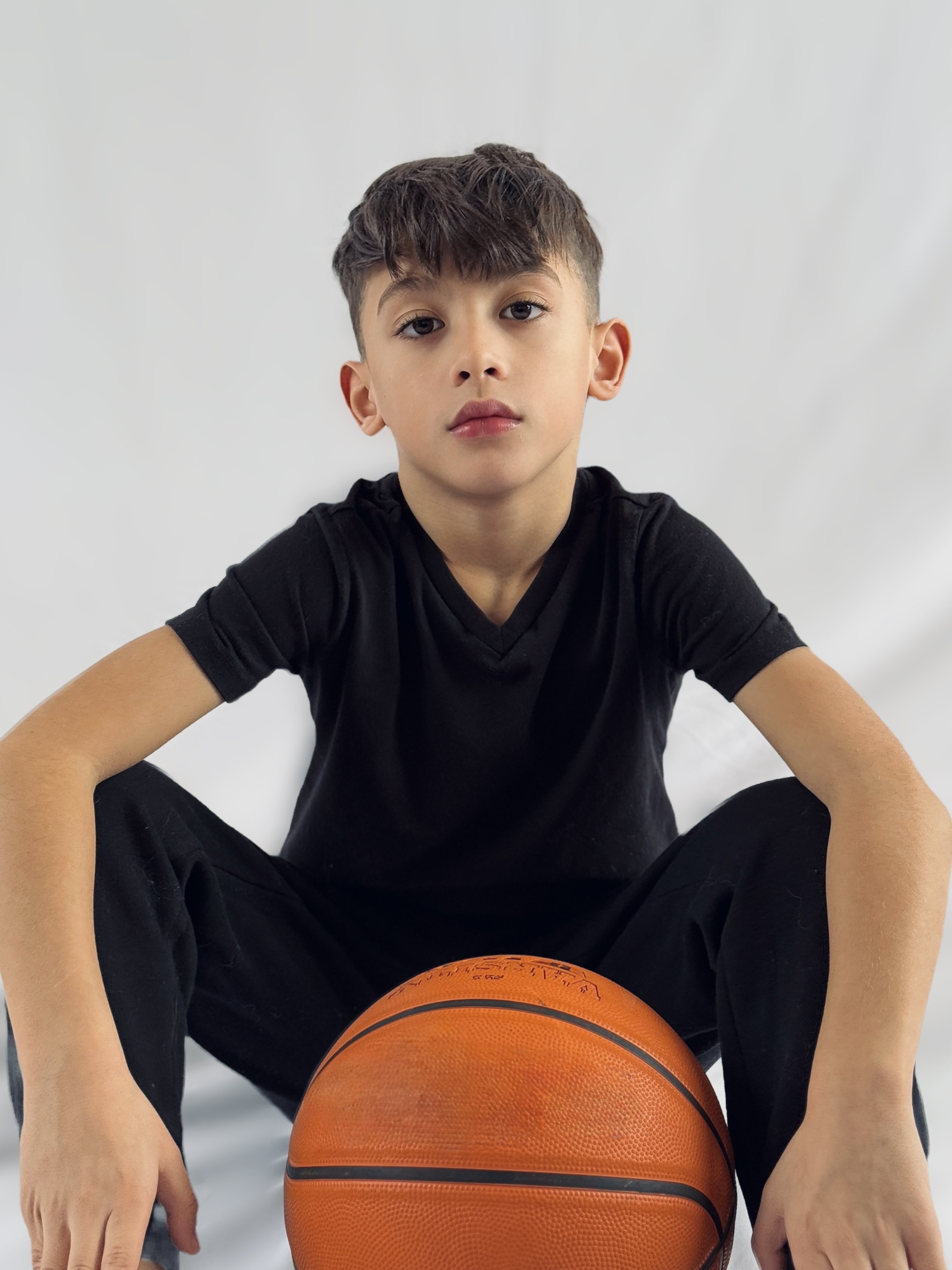 Young boy with short dark hair and brown eyes sitting down with a basketball between his legs, wearing a black shirt and black pants, against a plain white background.