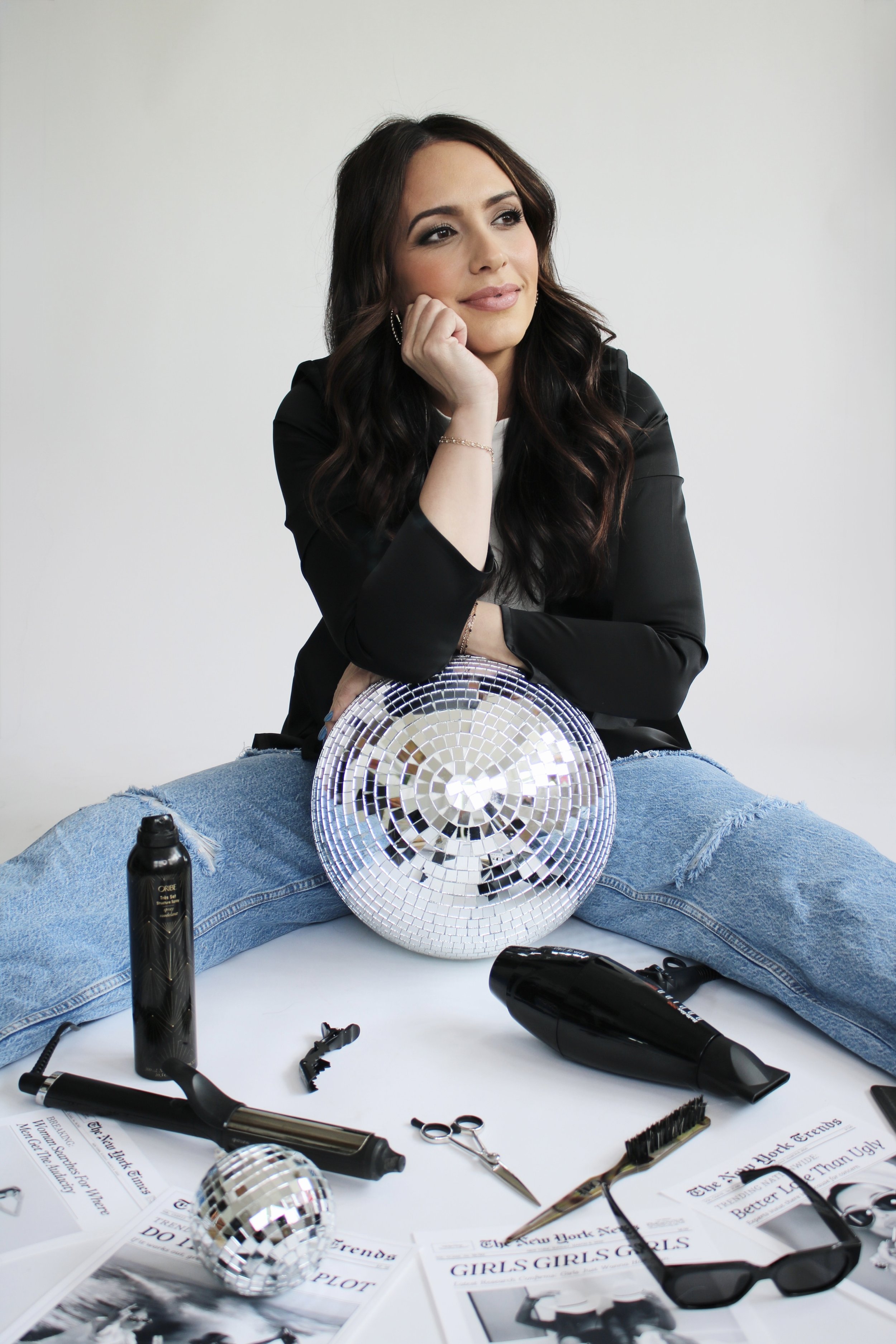 A woman sitting on the floor with her legs spread apart, surrounded by magazines, a disco ball, and hair styling tools including a hairdryer and curling iron, with a contemplative expression.