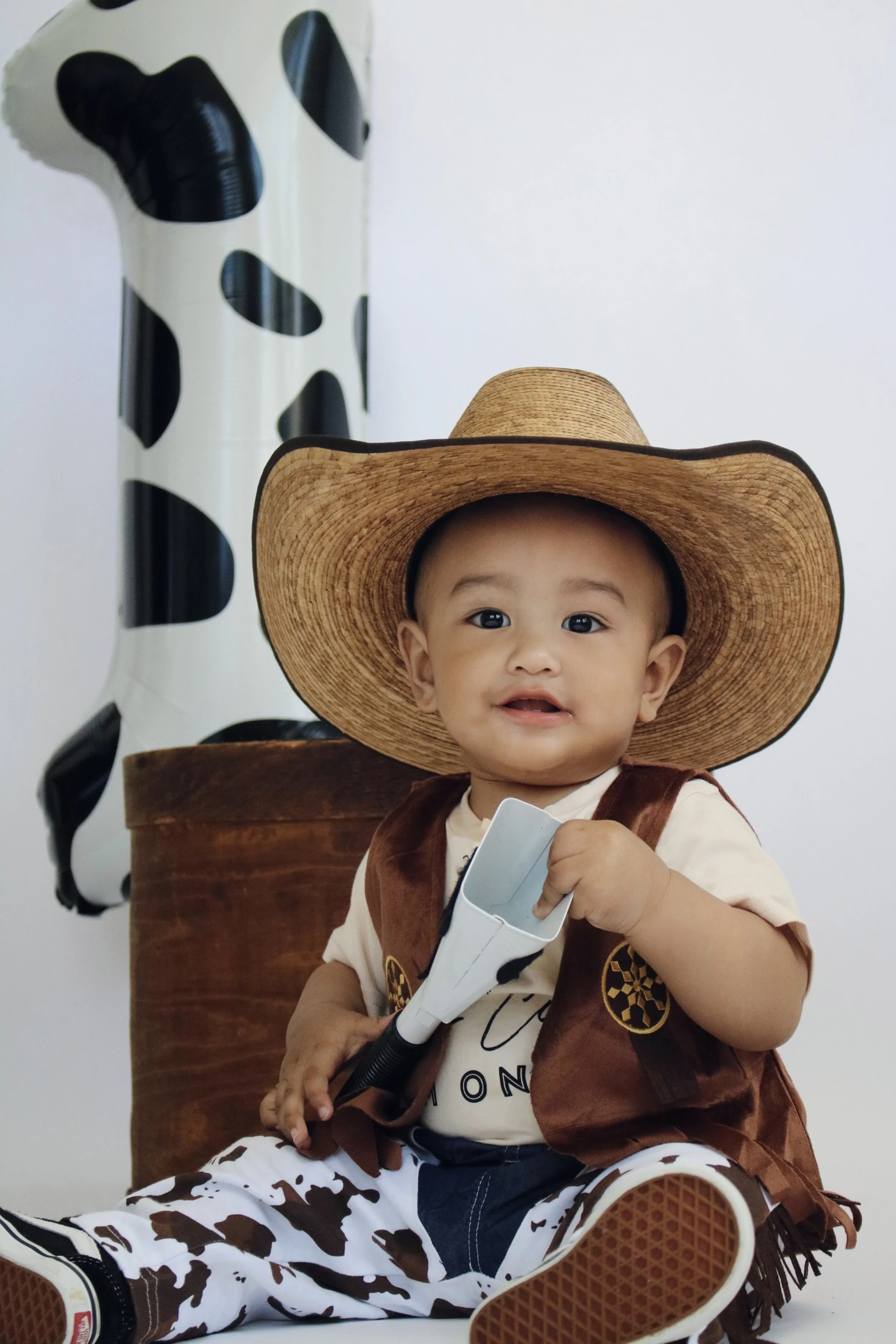 A smiling baby wearing a large brown cowboy hat, a tan shirt, a brown vest, and cow-print pants, sitting on the floor holding a toy gun, with a cow-print balloon and a wooden box in the background.