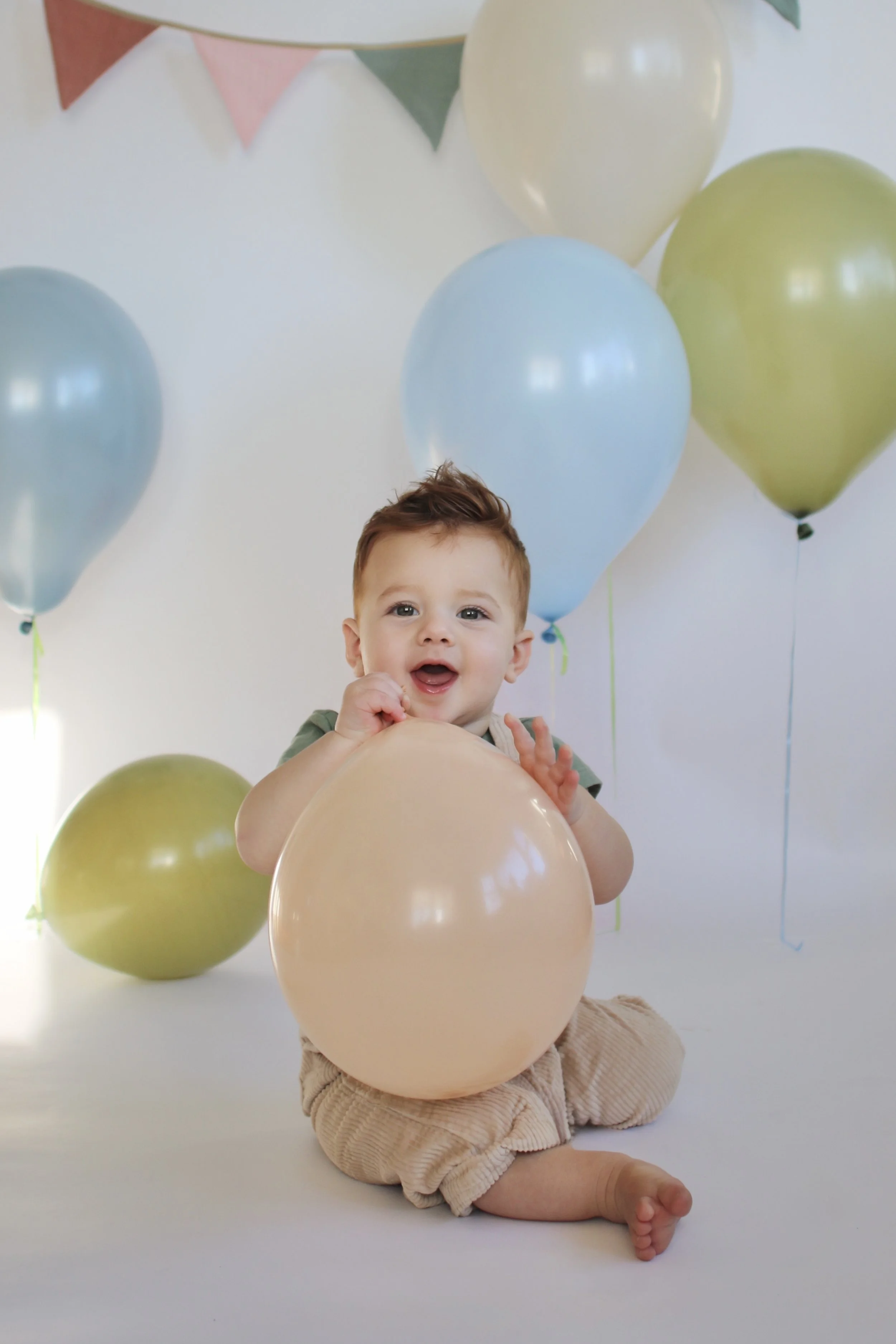 A happy baby sitting on the floor, holding a pink balloon, surrounded by pastel-colored balloons and decorative bunting, during a celebration or birthday party.