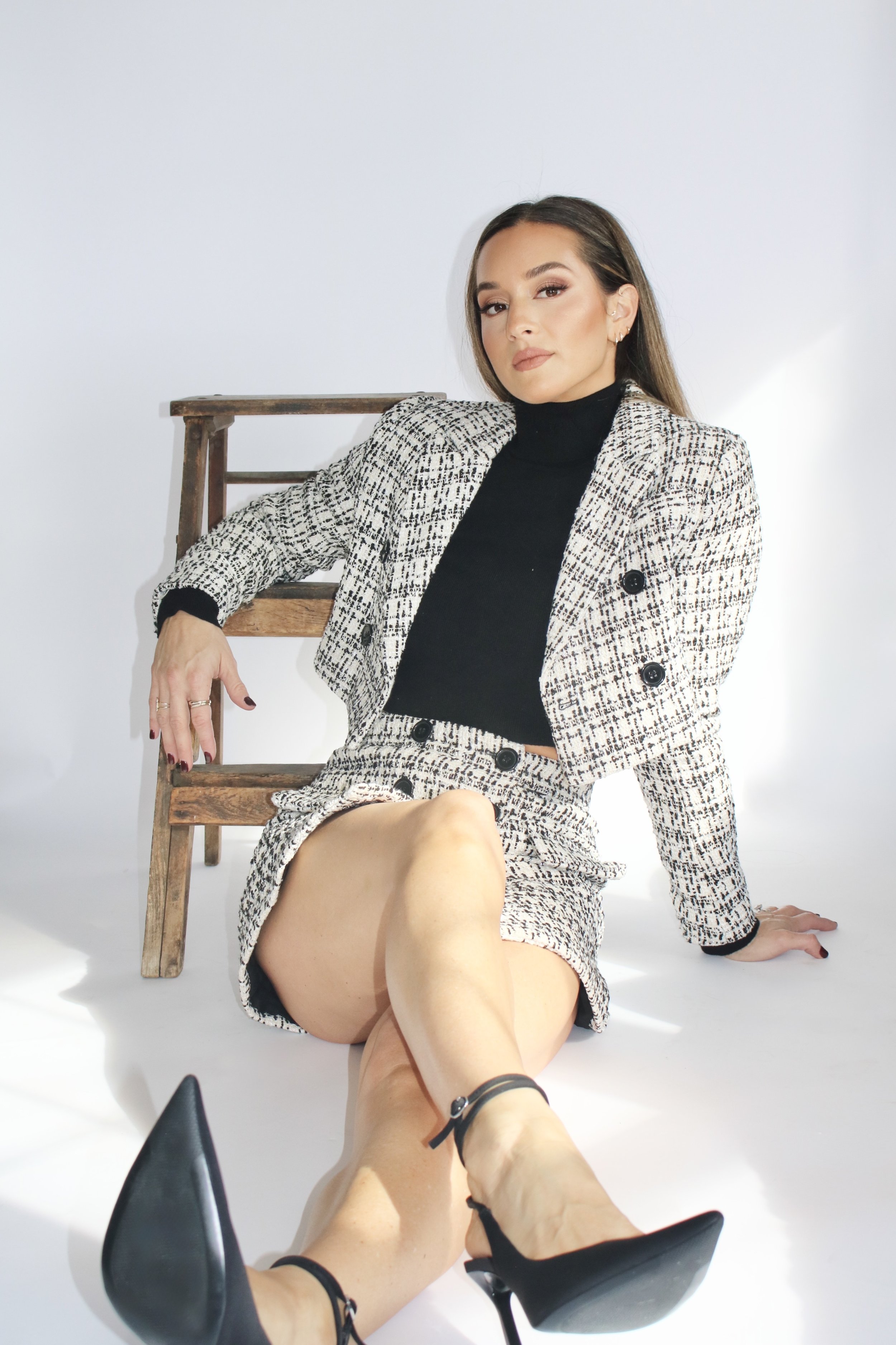 A woman sitting on the floor, leaning against a small wooden ladder, wearing a black turtleneck, patterned blazer and skirt, with black high heels.