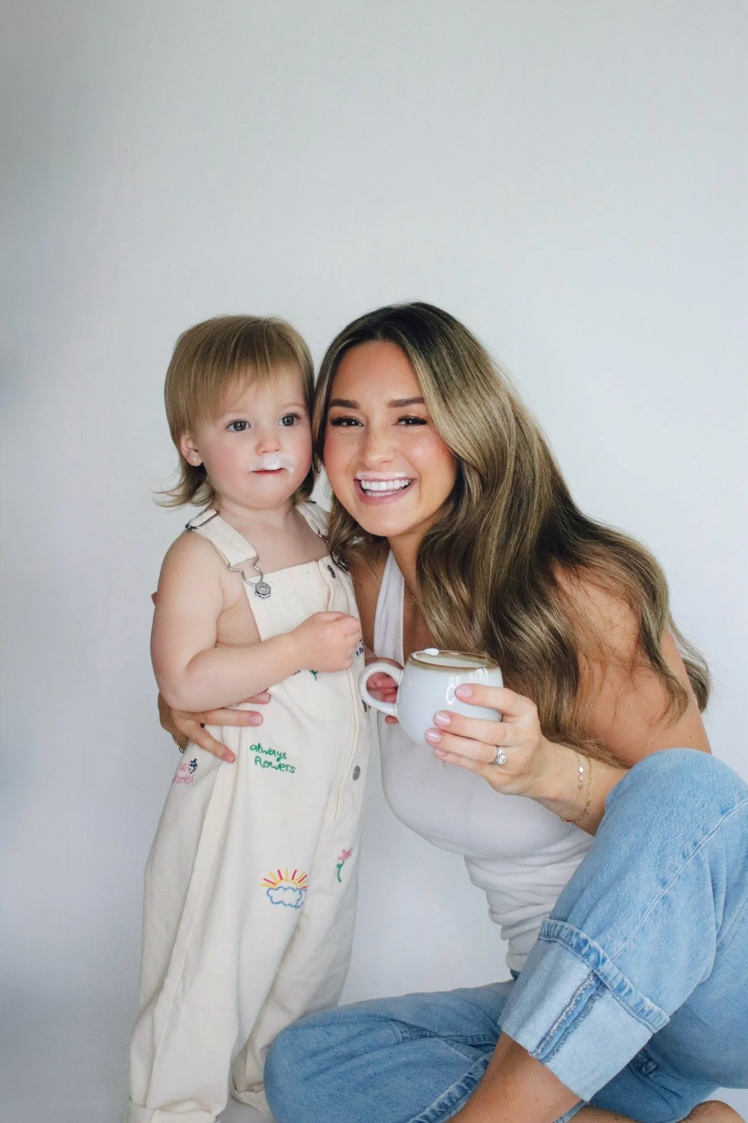 A woman smiling and holding a cup of coffee, sitting with a young girl with milk mustache, against a plain white wall.