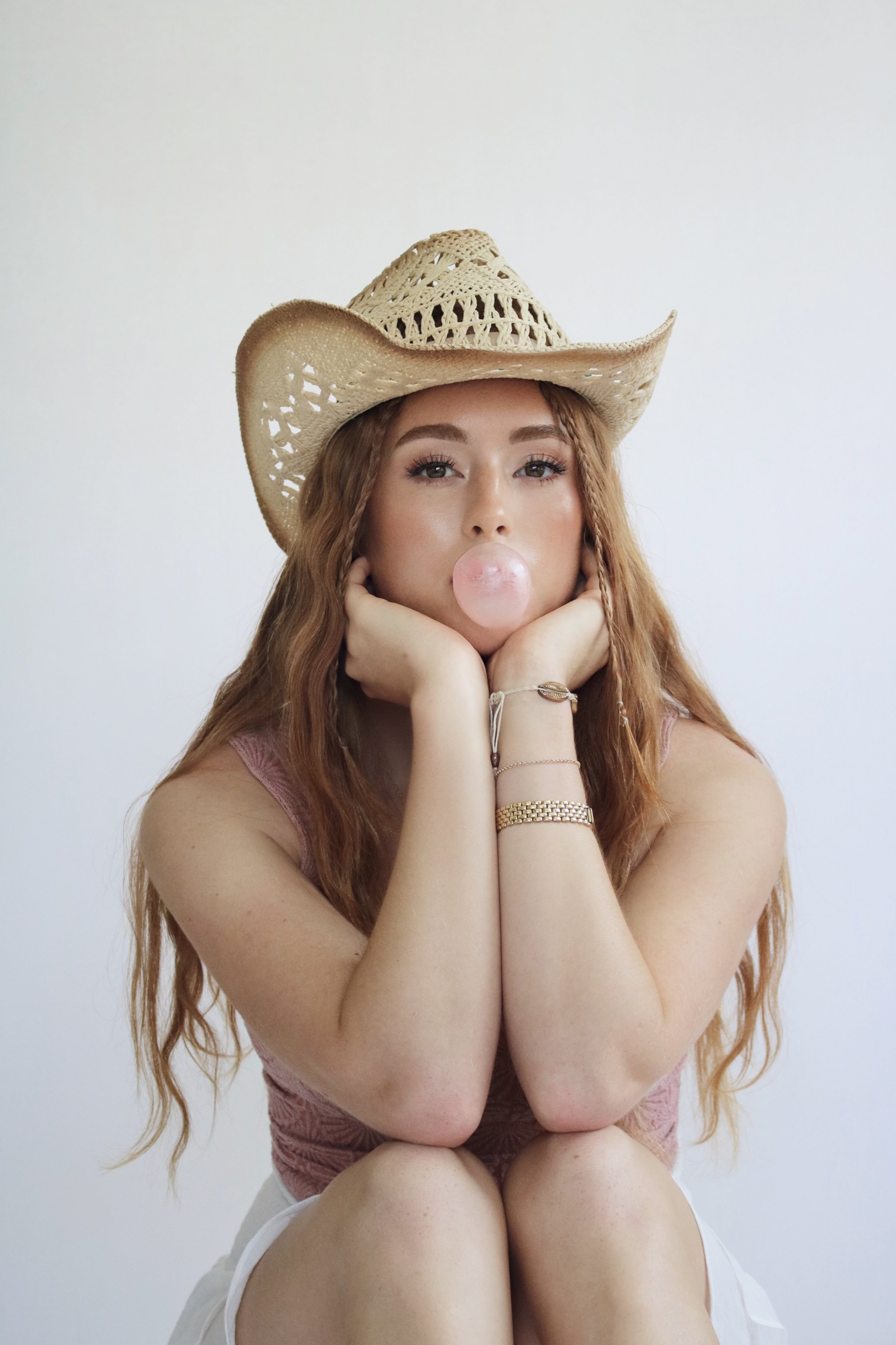 A young woman with long red hair, wearing a straw cowboy hat, is sitting with her chin resting on her hands. She is blowing a bubble gum bubble and dressed in a pink sleeveless top and white shorts, against a plain white background.