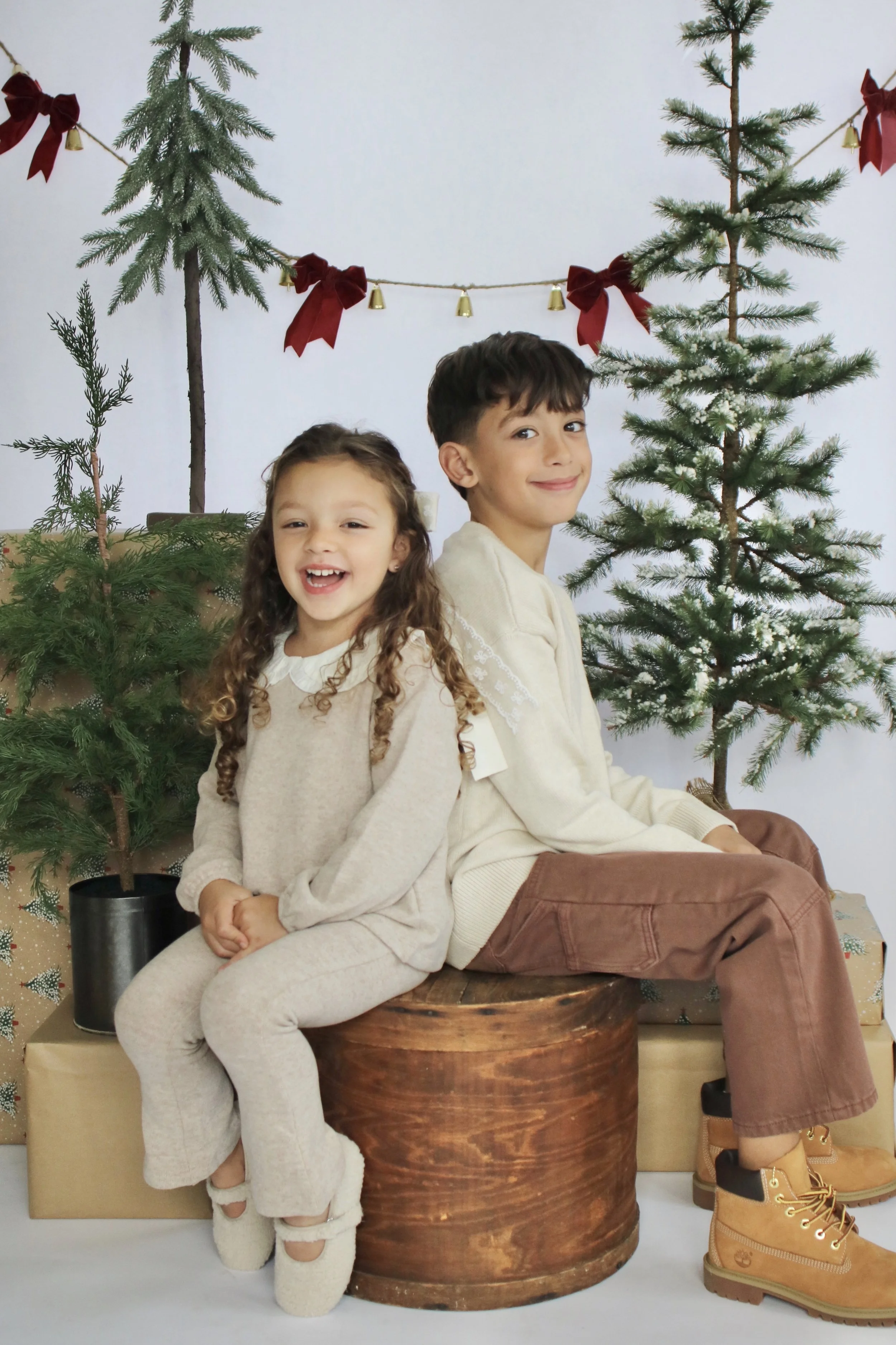 A girl and boy sitting back to back in front of a Christmas scene with small decorated trees, wrapped gifts, and holiday decorations.