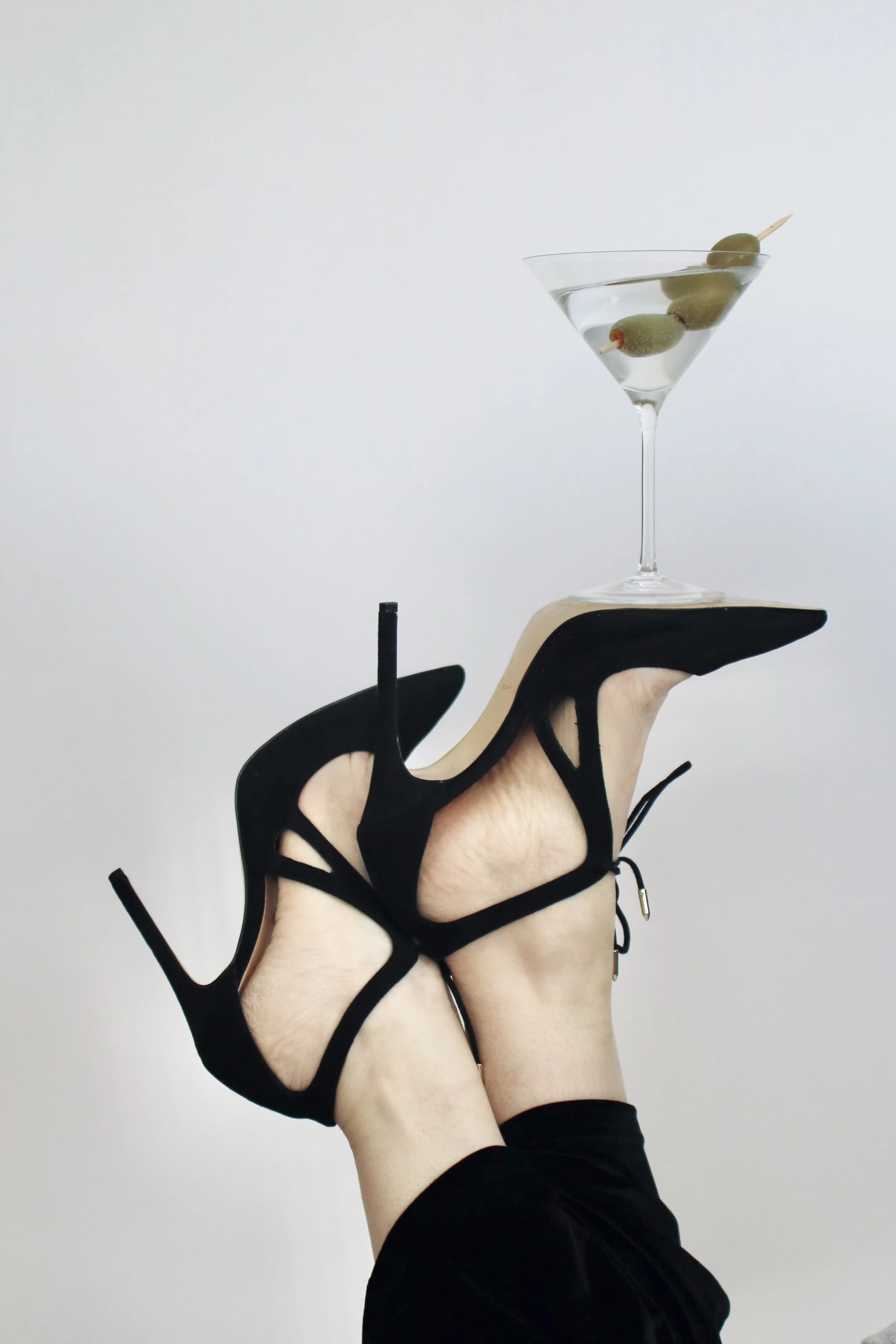 Person wearing black high heels with their legs raised, balancing a cocktail glass with olives on the heel of one shoe.
