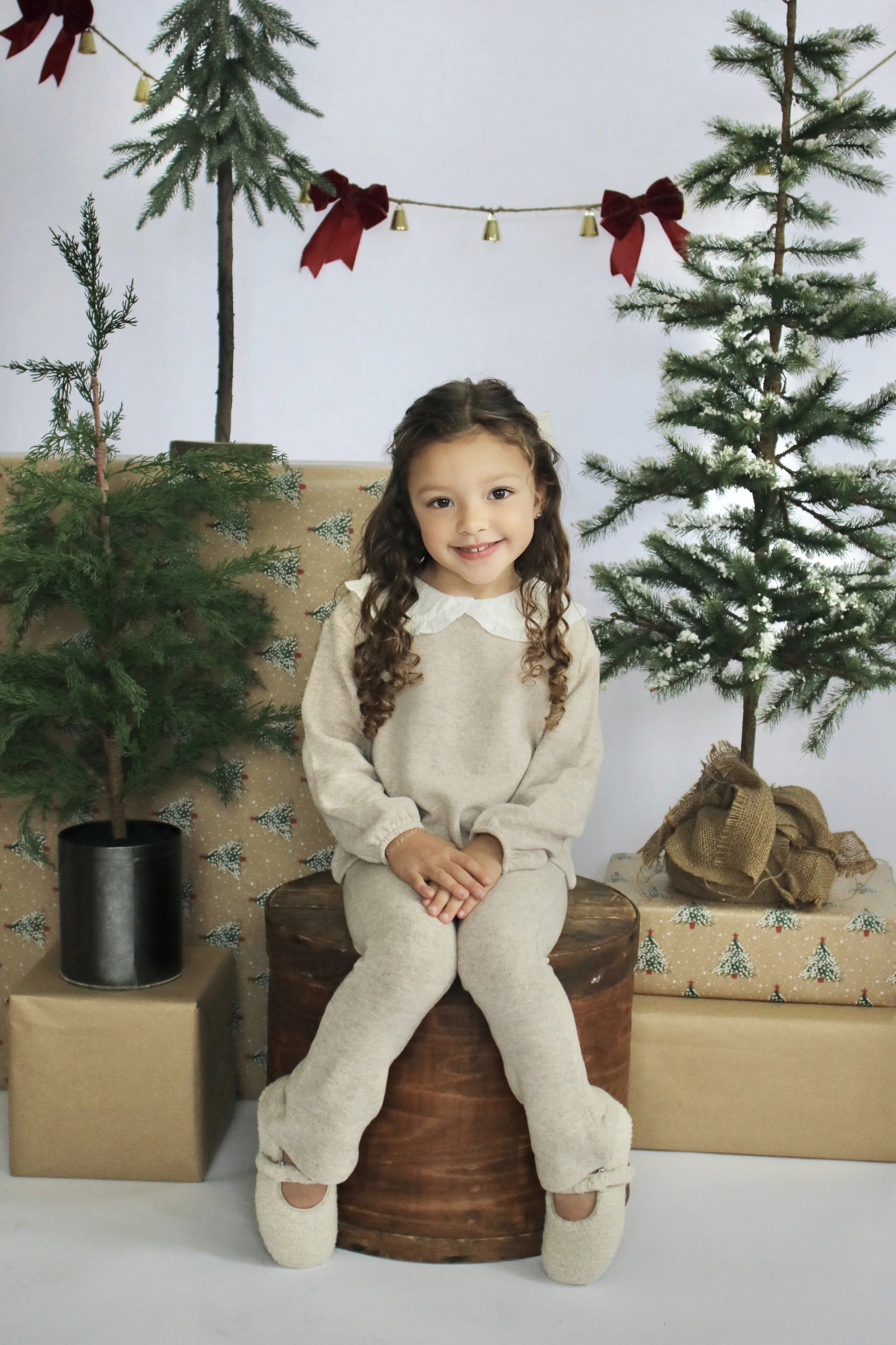 A young girl with long curly hair sitting on a wooden stump, surrounded by Christmas trees and wrapped presents, with festive decorations hanging above.