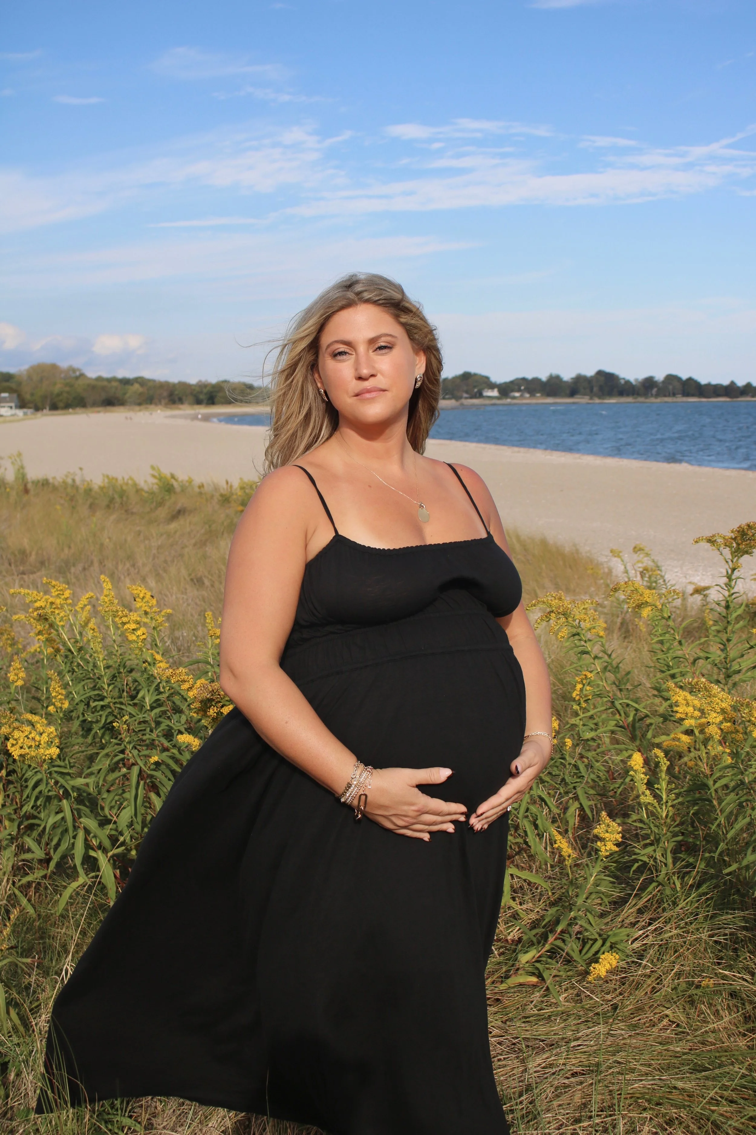 A pregnant woman in a black dress standing outdoors on a grassy beach with yellow flowers, holding her belly, near a body of water under a partly cloudy sky.