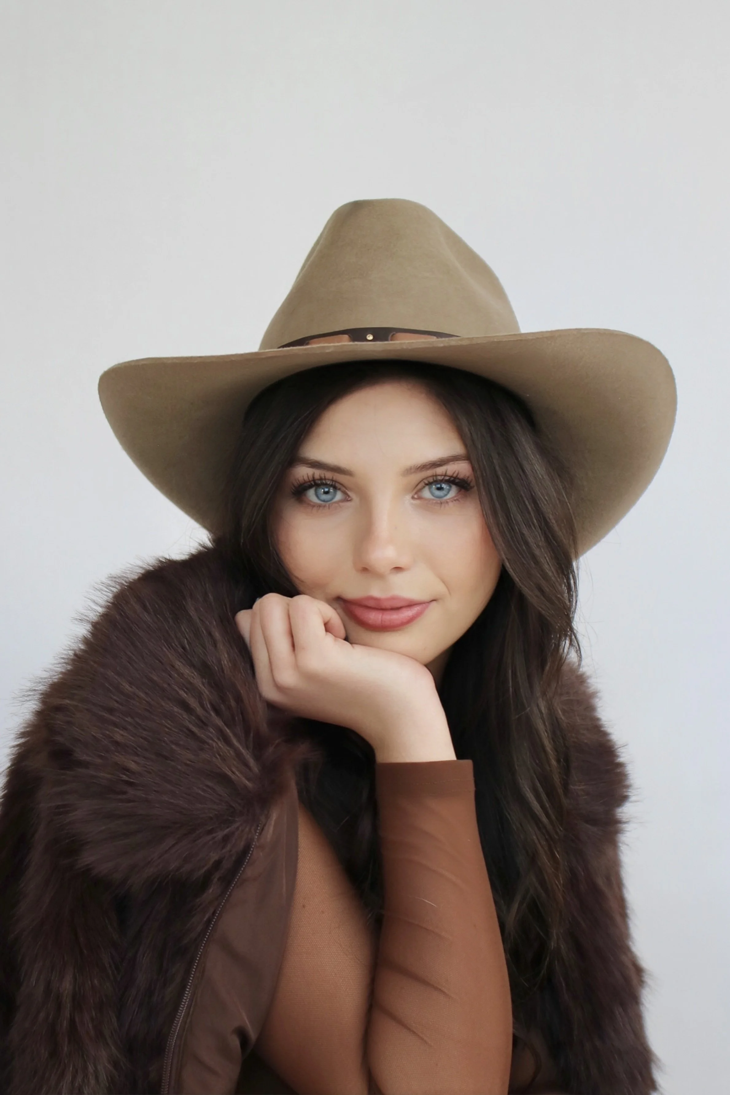 A woman with long dark hair, blue eyes, and a light complexion, wearing a large beige cowboy hat, a brown fur coat, and sheer brown sleeves, resting her chin on her hand and looking at the camera with a slight smile.