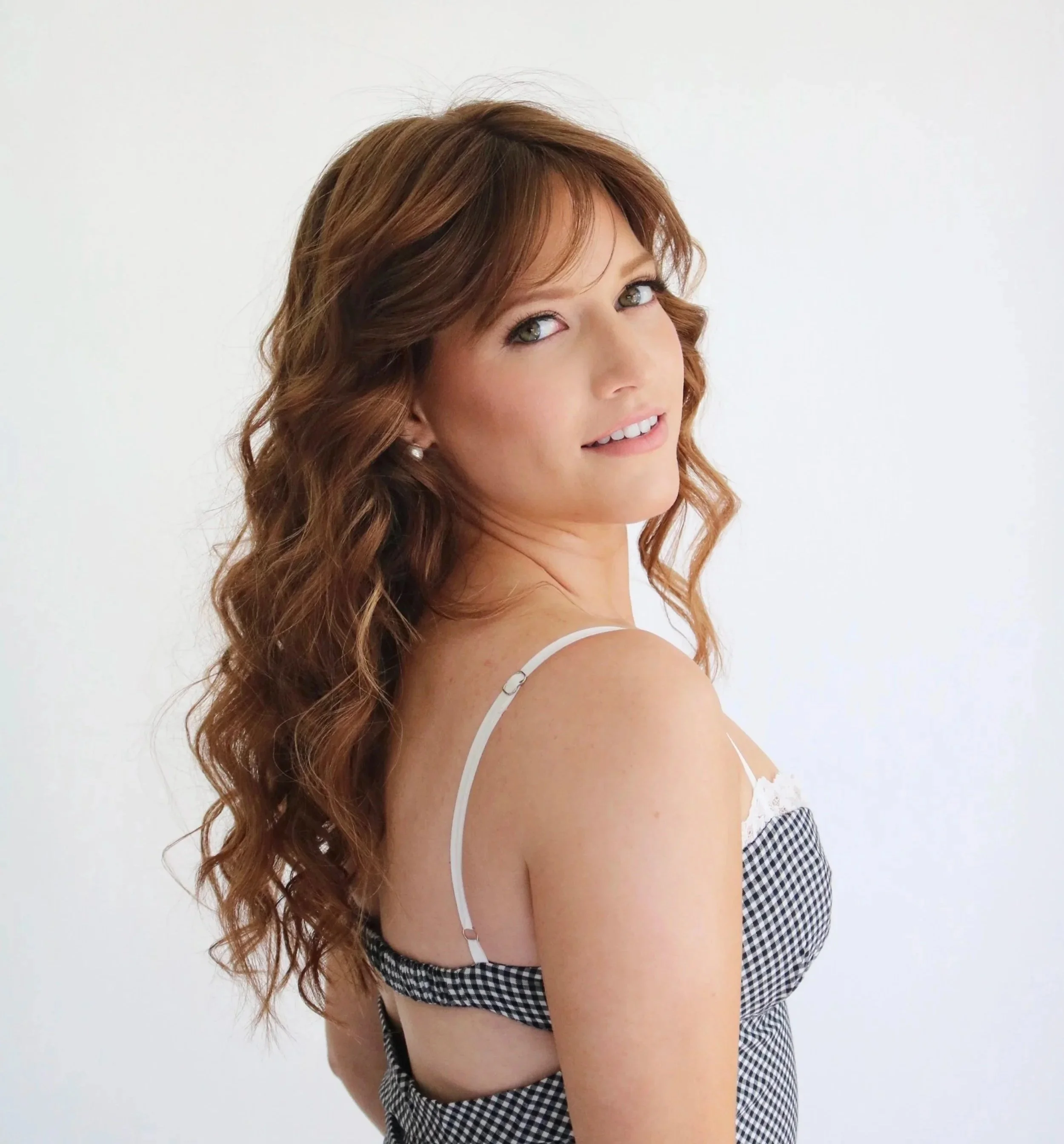 A woman with long, curly auburn hair and light makeup, wearing a sleeveless checkered dress with white straps, posing against a plain white background.