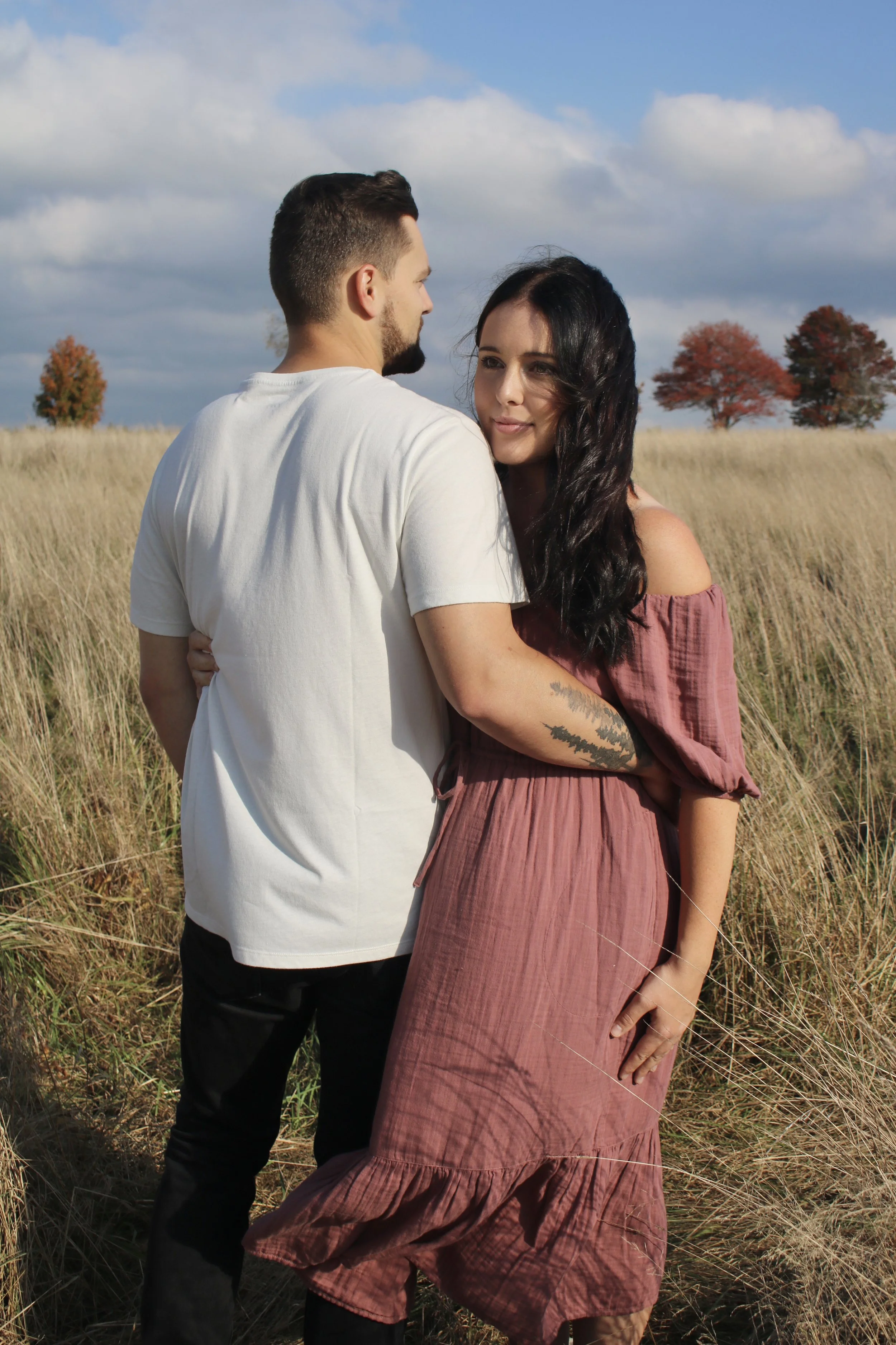 A man and woman standing close together in a grassy field, with trees in the background and a partly cloudy sky.