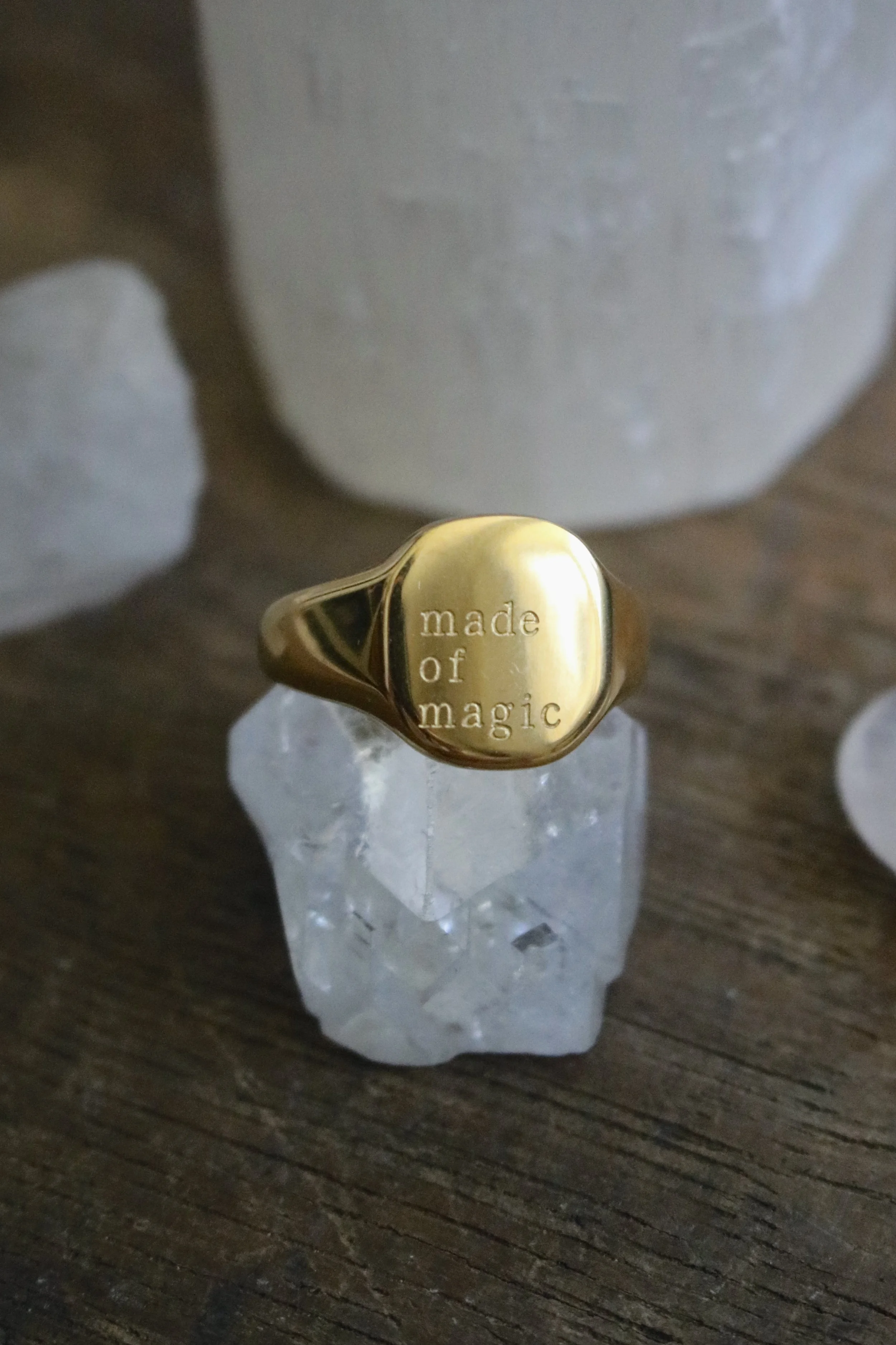 A gold ring engraved with the words "made of magic" resting on a clear, faceted crystal placed on a wooden surface.