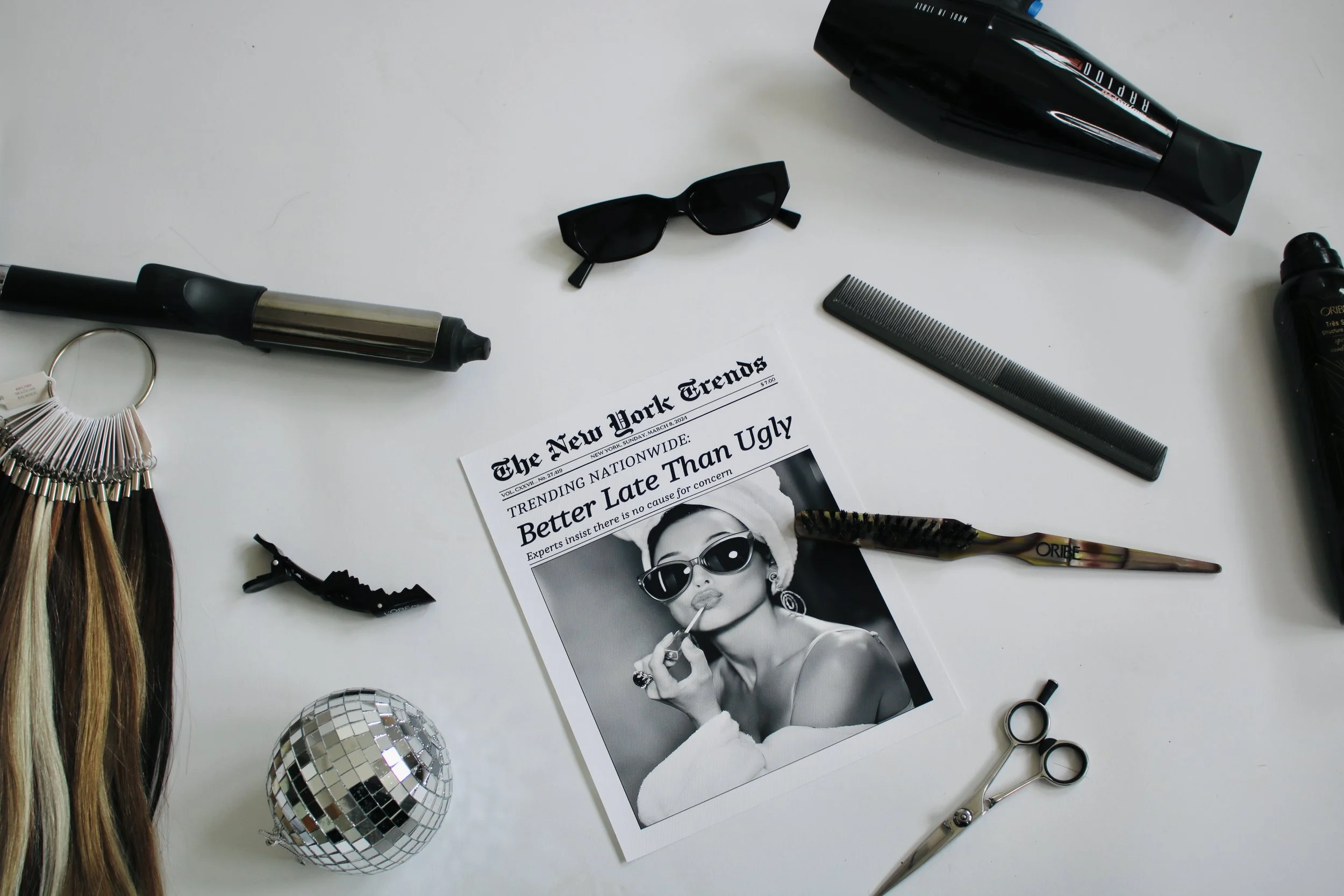 Hair styling tools, sunglasses, a comb, scissors, a doll headbrush, a small disco ball, and a newspaper with a black-and-white photo of a woman wearing sunglasses and makeup on a white surface.
