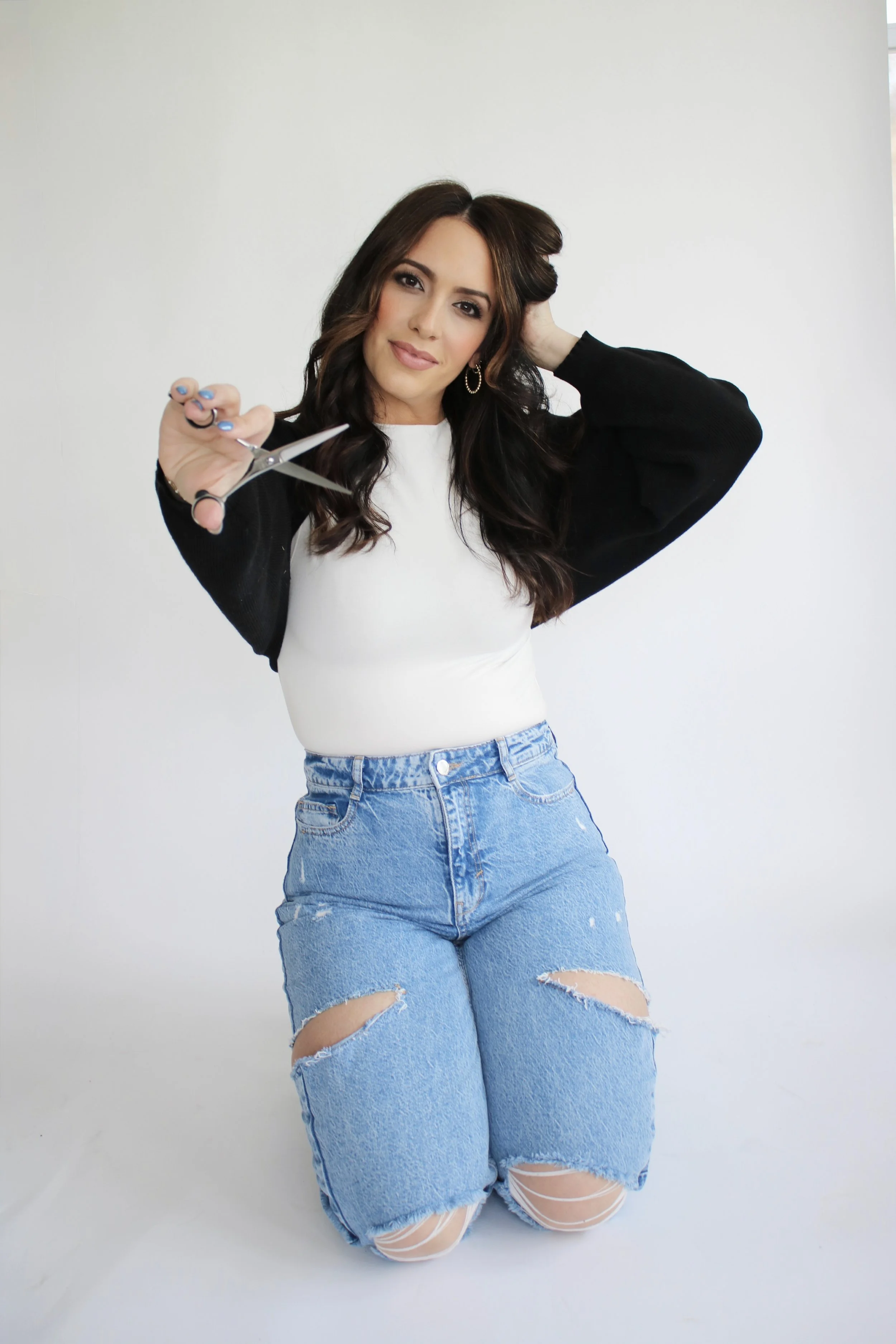 A woman with long dark hair wearing ripped blue jeans, a white top, and a black cardigan. She is kneeling and holding a pair of scissors towards the camera, with a slight smile on her face.