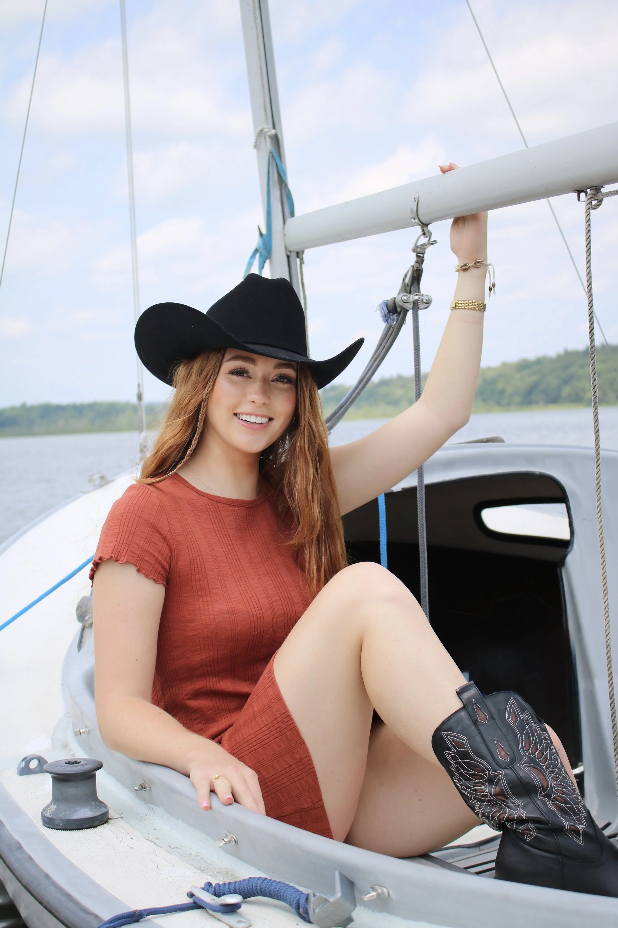 A young woman wearing a black cowboy hat and a rust-colored dress sitting on a sailboat, holding onto the sailboat mast, smiling at the camera, with a body of water and a green shoreline in the background.