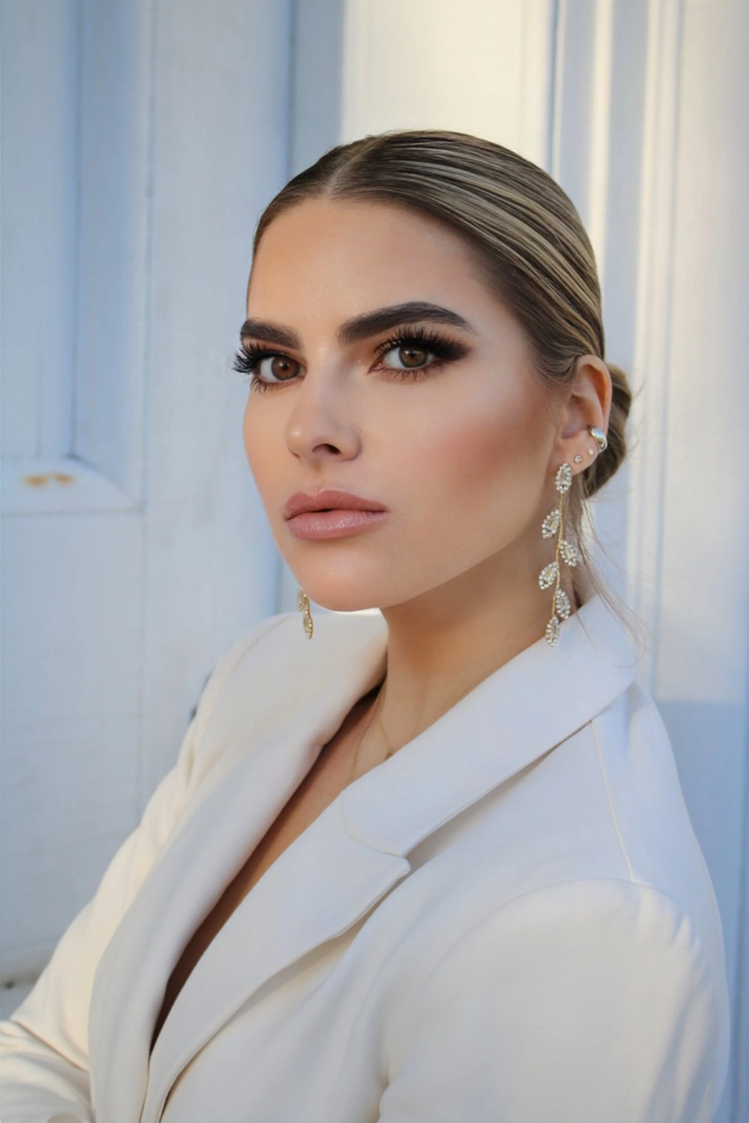 A woman with styled hair, noticeable makeup, and dangling leaf-shaped earrings posing against a light-colored background.