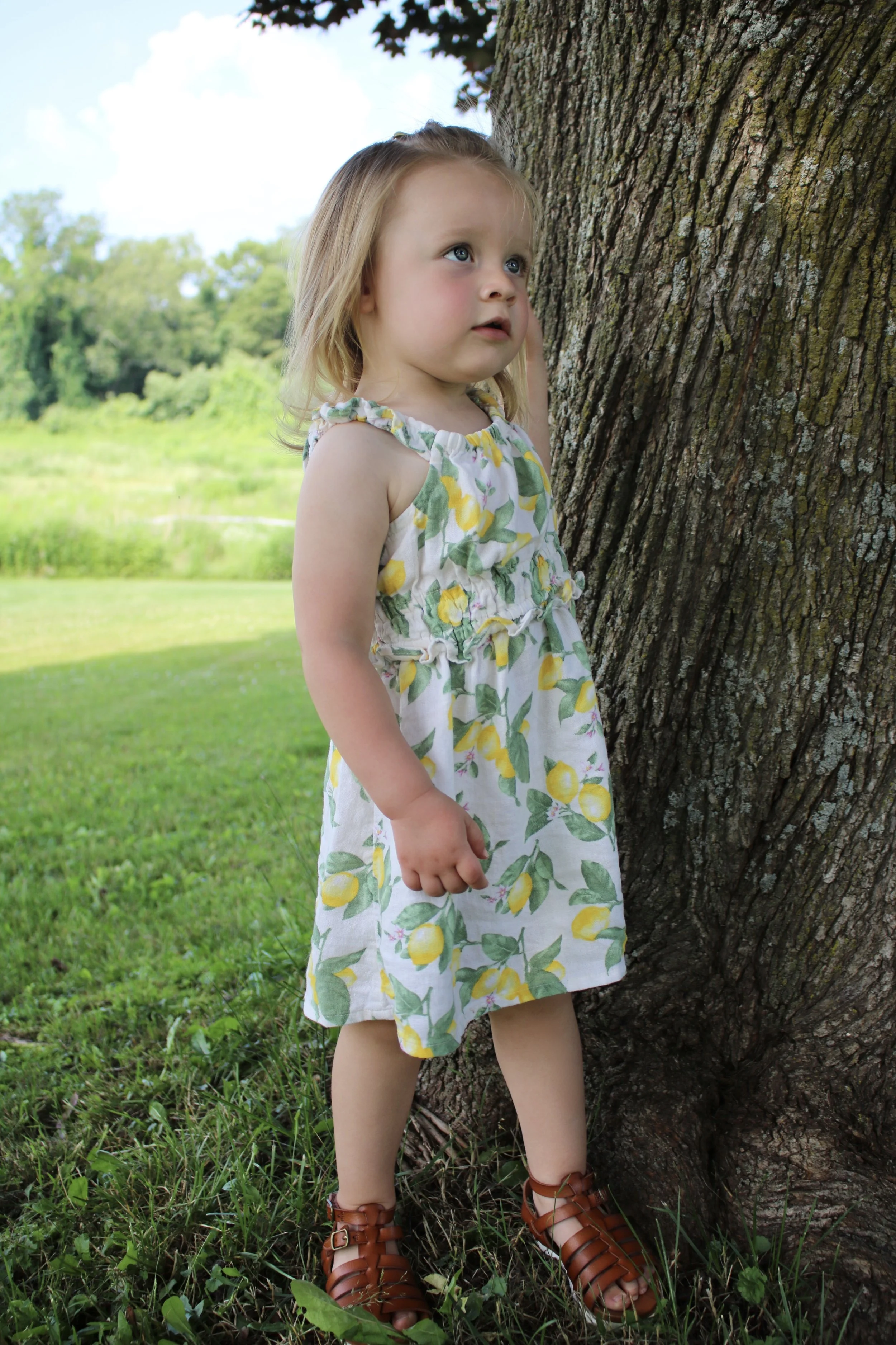 A young girl with blonde hair and blue eyes standing next to a large tree trunk in a grassy outdoor setting, wearing a lemon-patterned dress and brown sandals, looking off into the distance.
