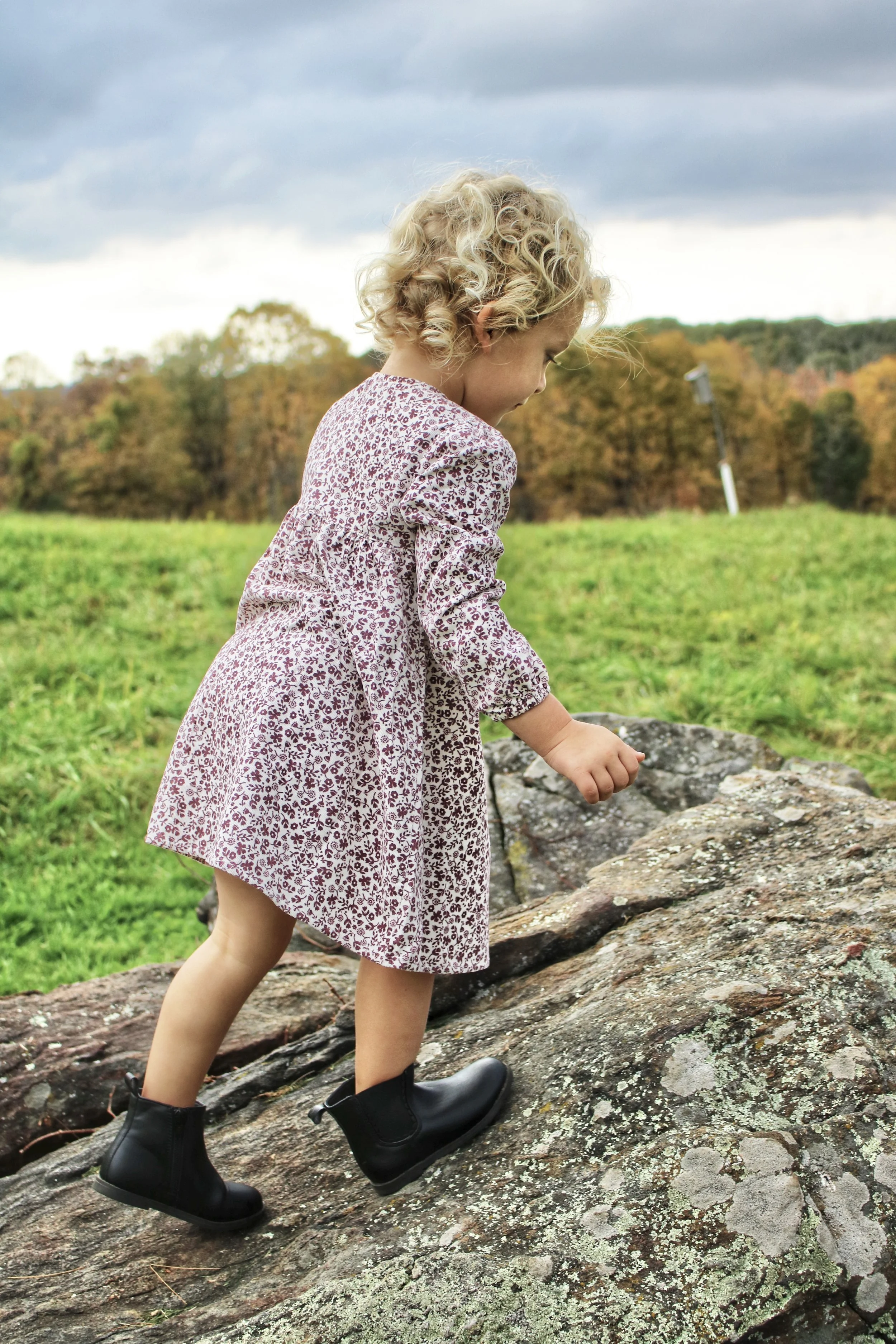 A young girl with curly blonde hair wearing a floral dress and black boots is climbing a large rock outdoors on a cloudy day, with a grassy field and trees in the background.