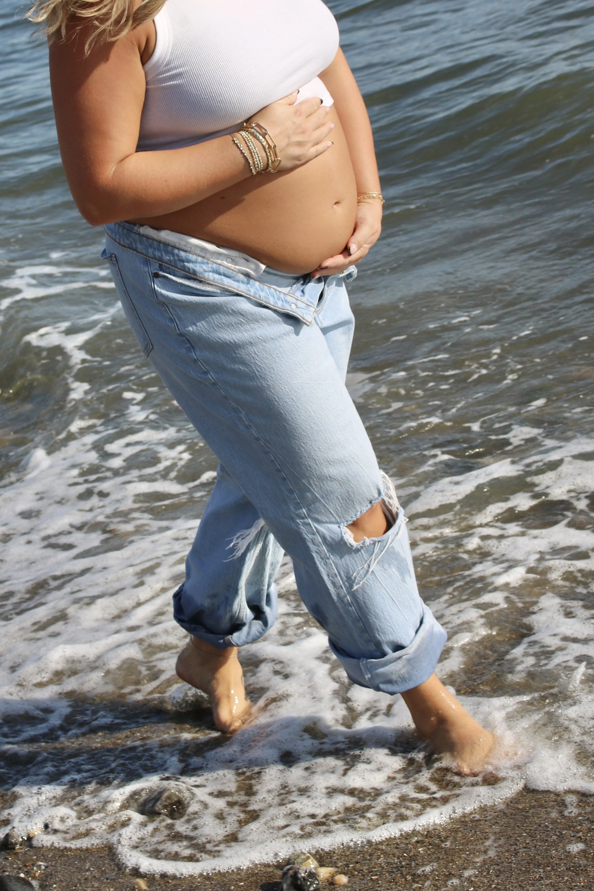 A pregnant woman walking in the ocean, wearing ripped jeans and a white sleeveless top, with her hand on her belly.