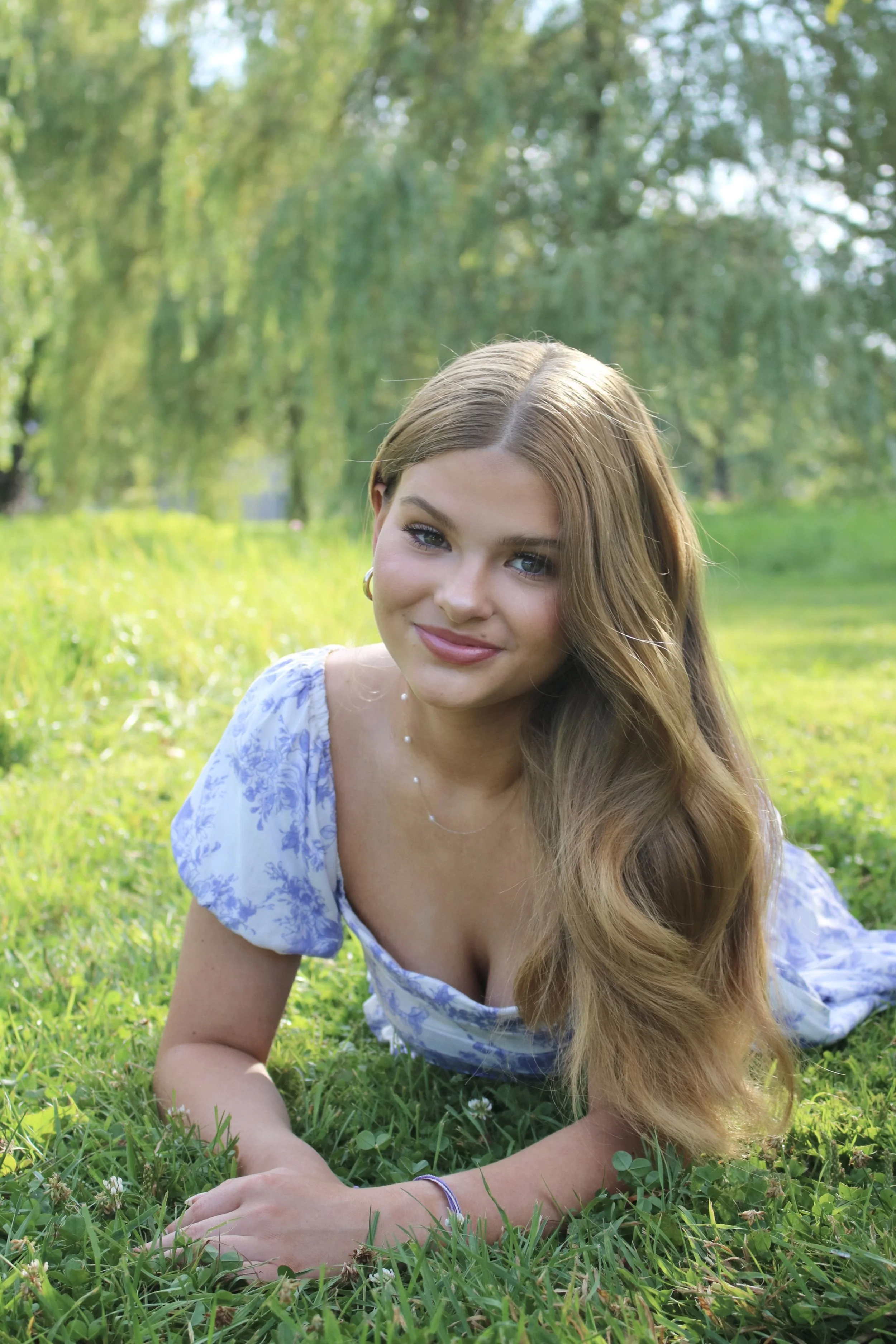 A young woman with long, wavy hair lying on grass in a park during daytime, smiling at the camera with a background of trees and sunlight.