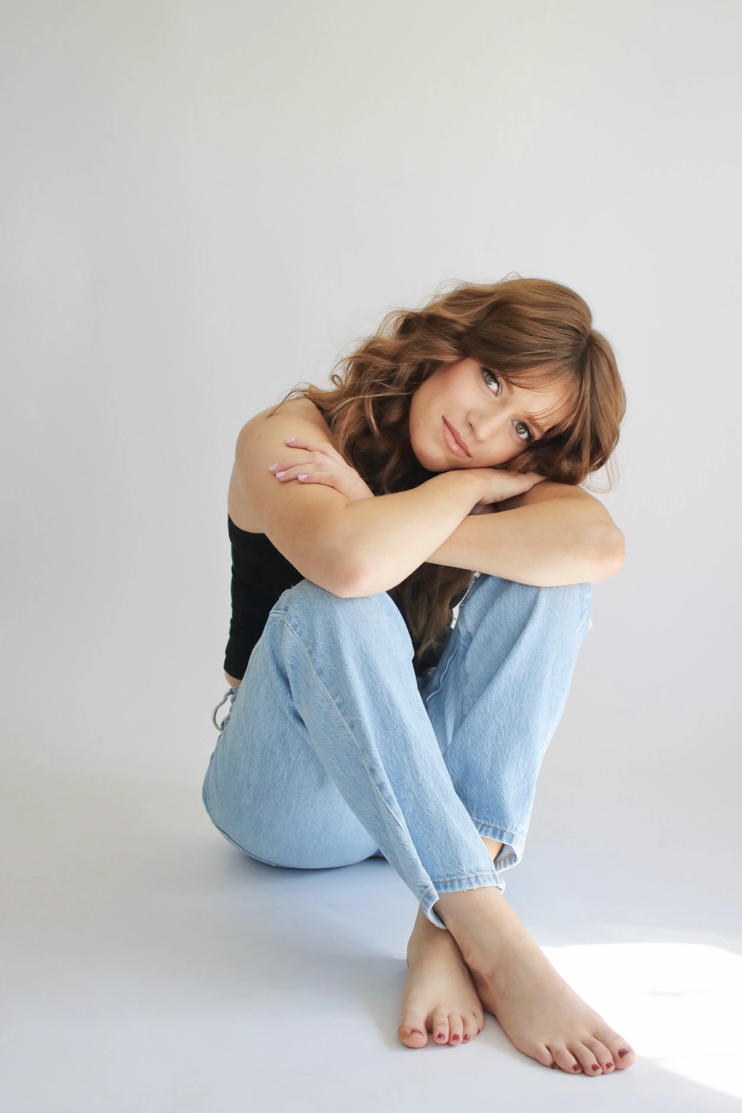 A woman with long wavy brown hair sitting on the floor with her knees drawn up, wearing a black tank top and light blue jeans. She rests her chin on her crossed arms and looks into the camera with a gentle expression, against a plain white background