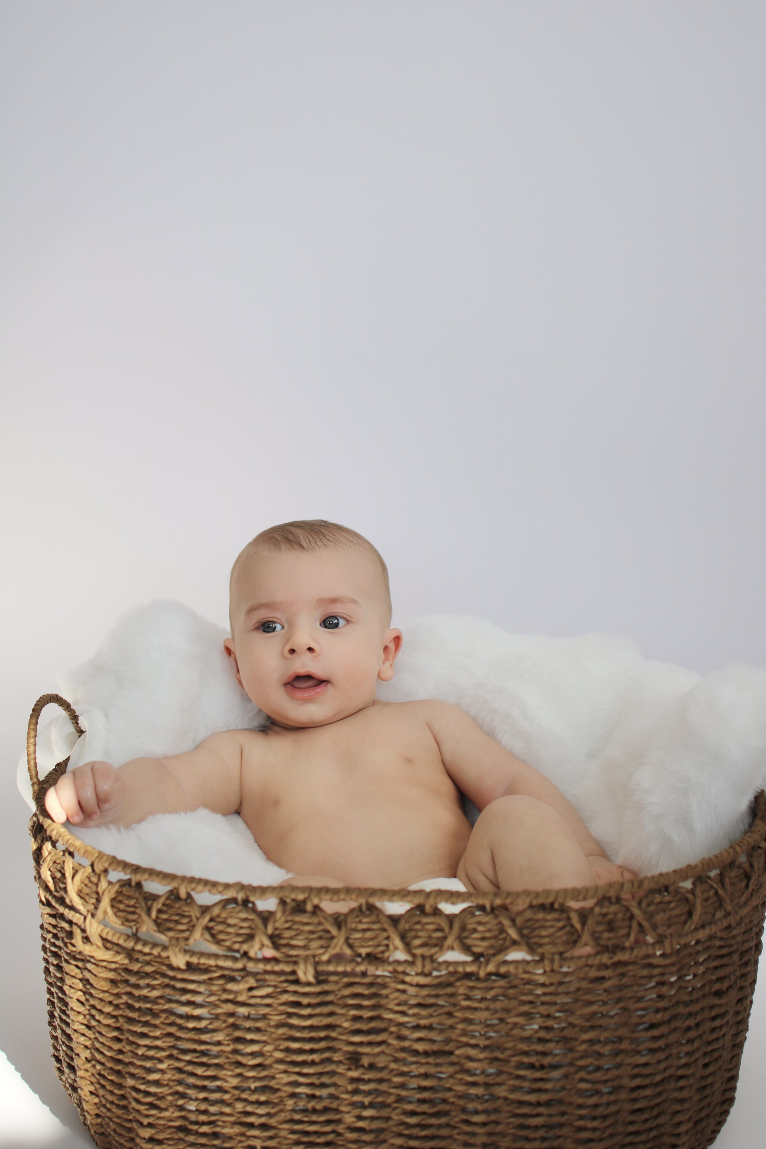 A baby with light skin, short light brown hair, and blue eyes, sitting in a brown woven basket lined with soft white fabric, against a plain white background.