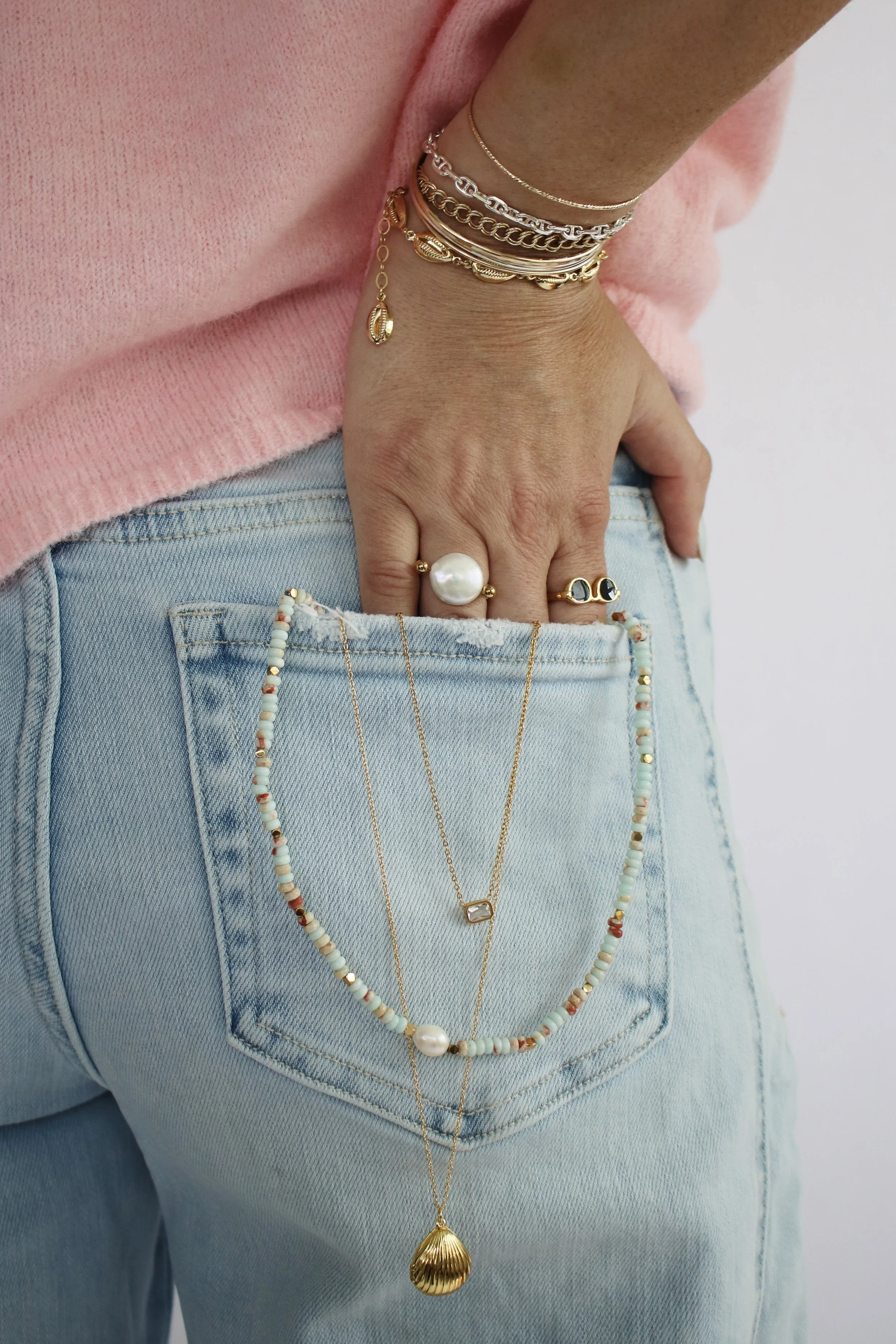 Person wearing multiple layered necklaces, several gold and silver bracelets, a large pearl ring, a gold ring with dark stones, and has jewelry draped over the back pocket of light-wash jeans while resting their hand on their hip.