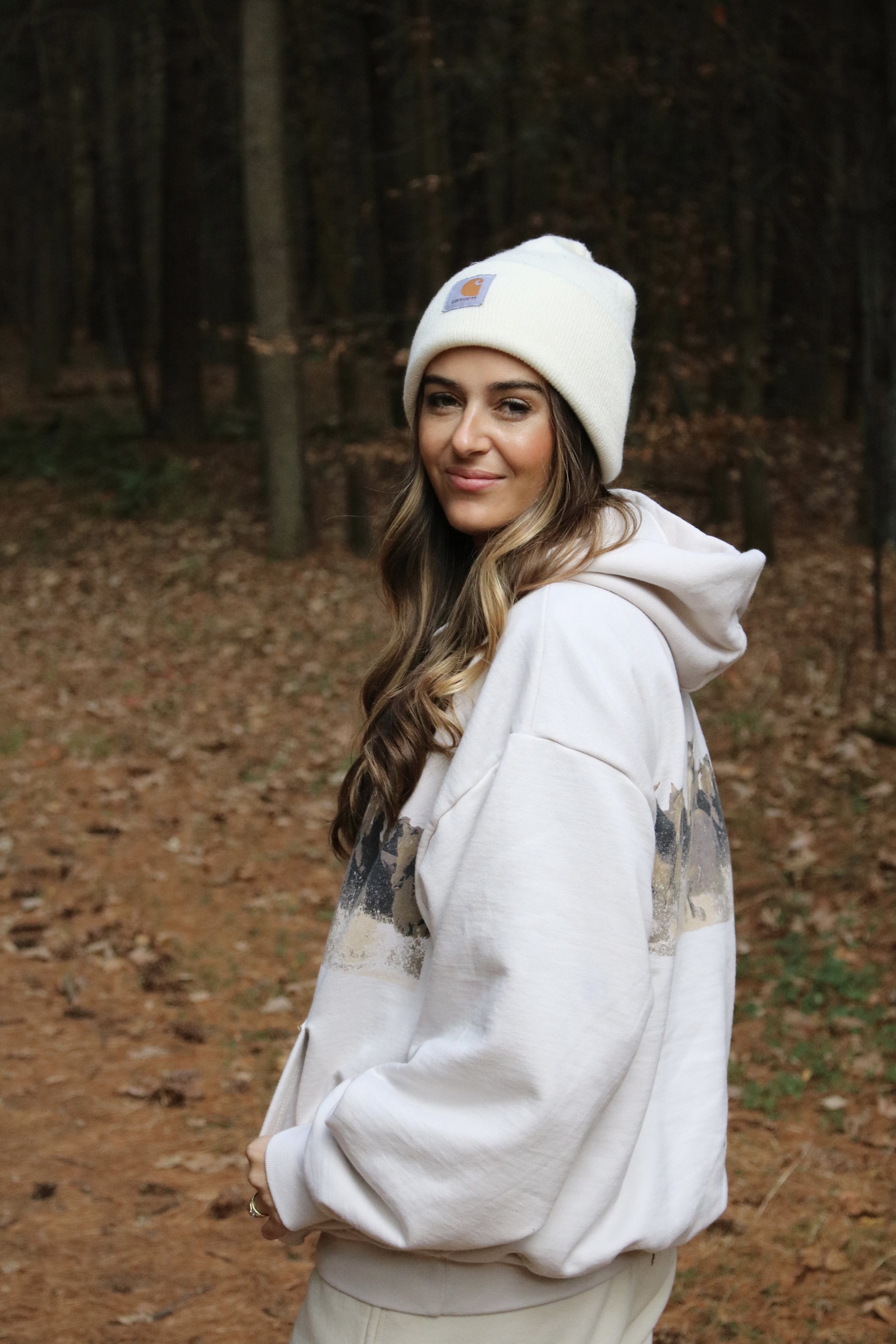 A young woman with long wavy hair wearing a white hoodie and a white beanie, standing in a wooded area with trees and fallen leaves.