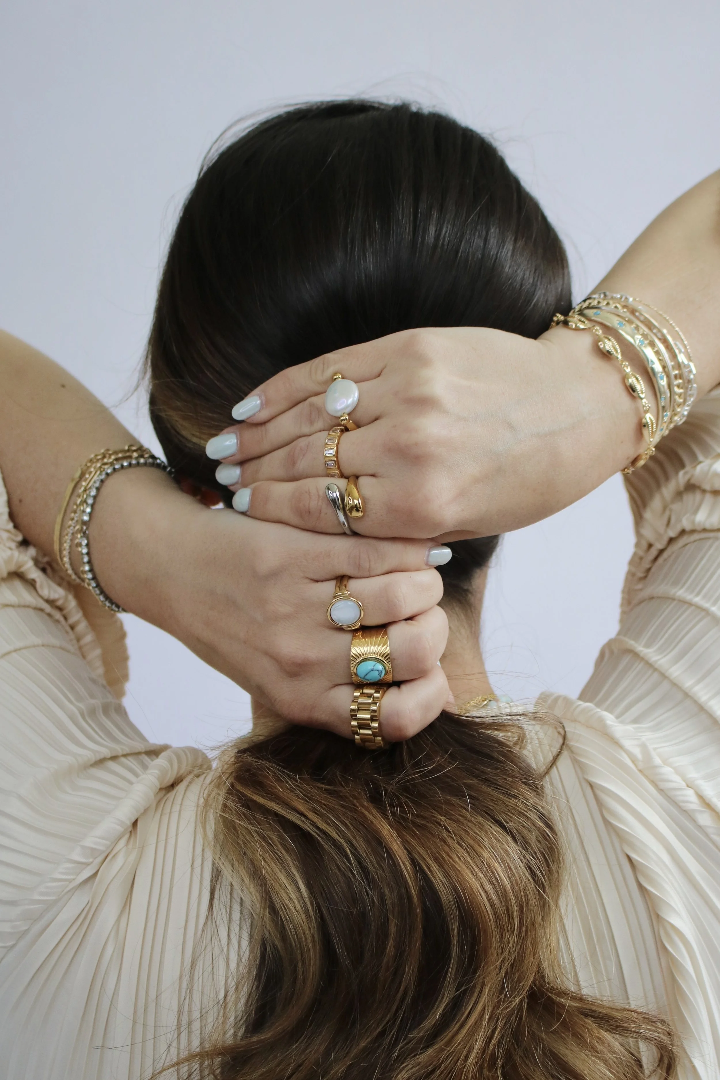A woman with brown hair styled in loose waves, wearing multiple gold and silver rings and jewelry on her hands and wrists, crossing her arms behind her head, revealing her nails painted light blue, and dressed in a beige, pleated top.