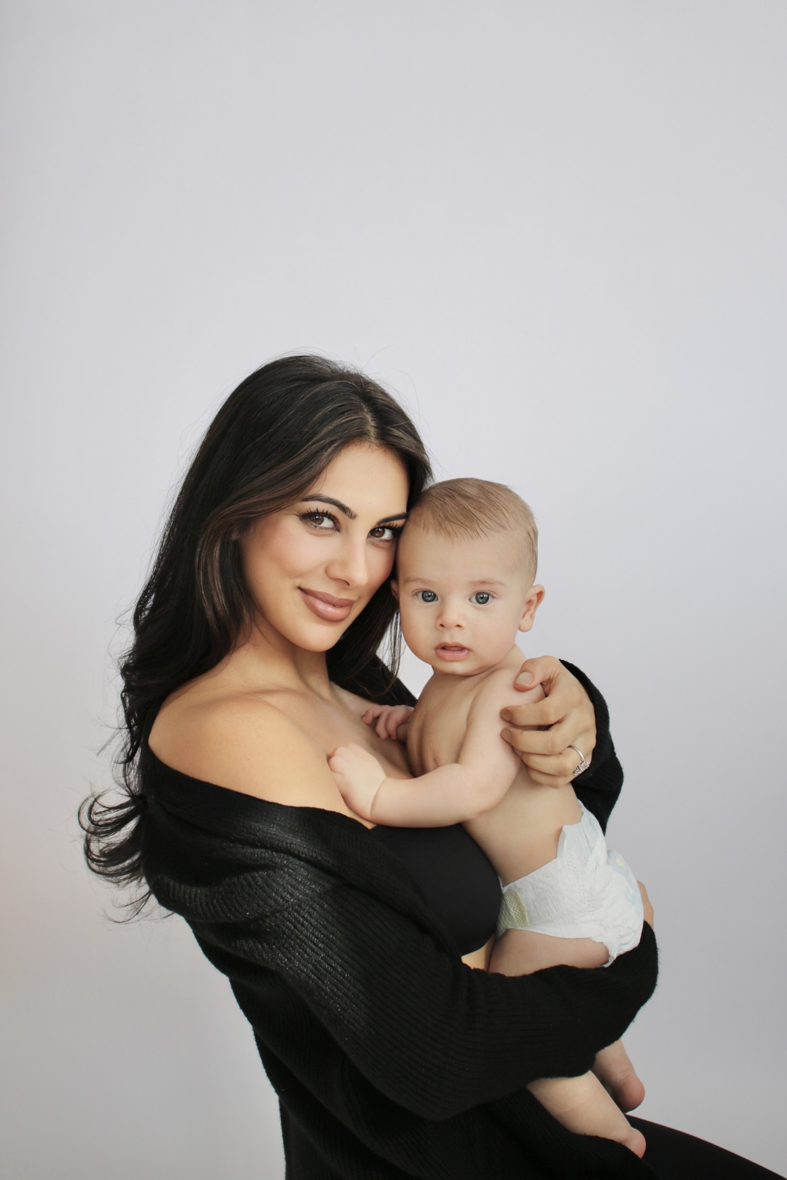 A woman with long dark hair holding a baby with blue eyes against a plain white background.