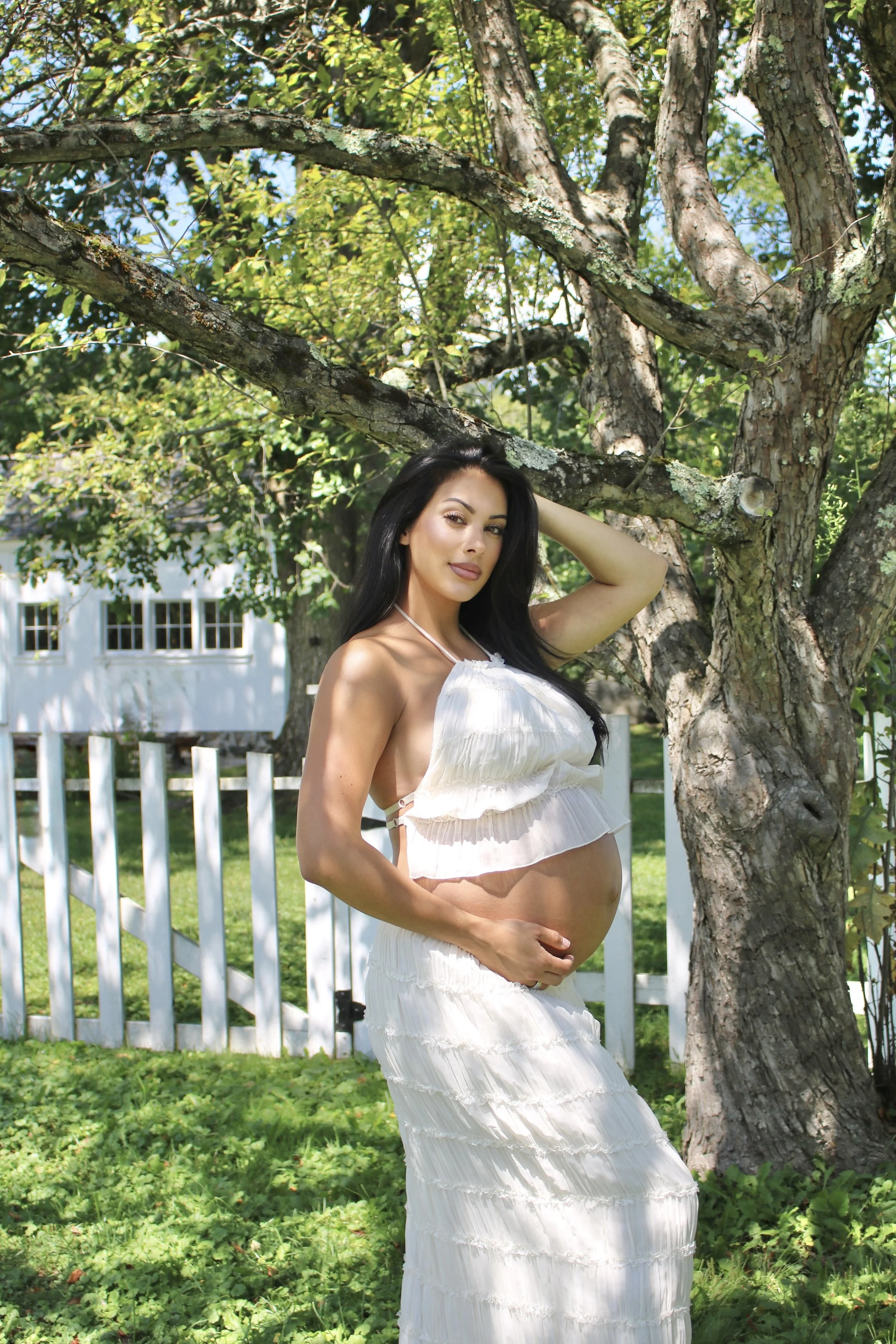 A pregnant woman stands outdoors in front of a tree, holding her belly with one hand and raising the other hand to touch her hair. She is wearing a white, ruffled, two-piece outfit and has long black hair.