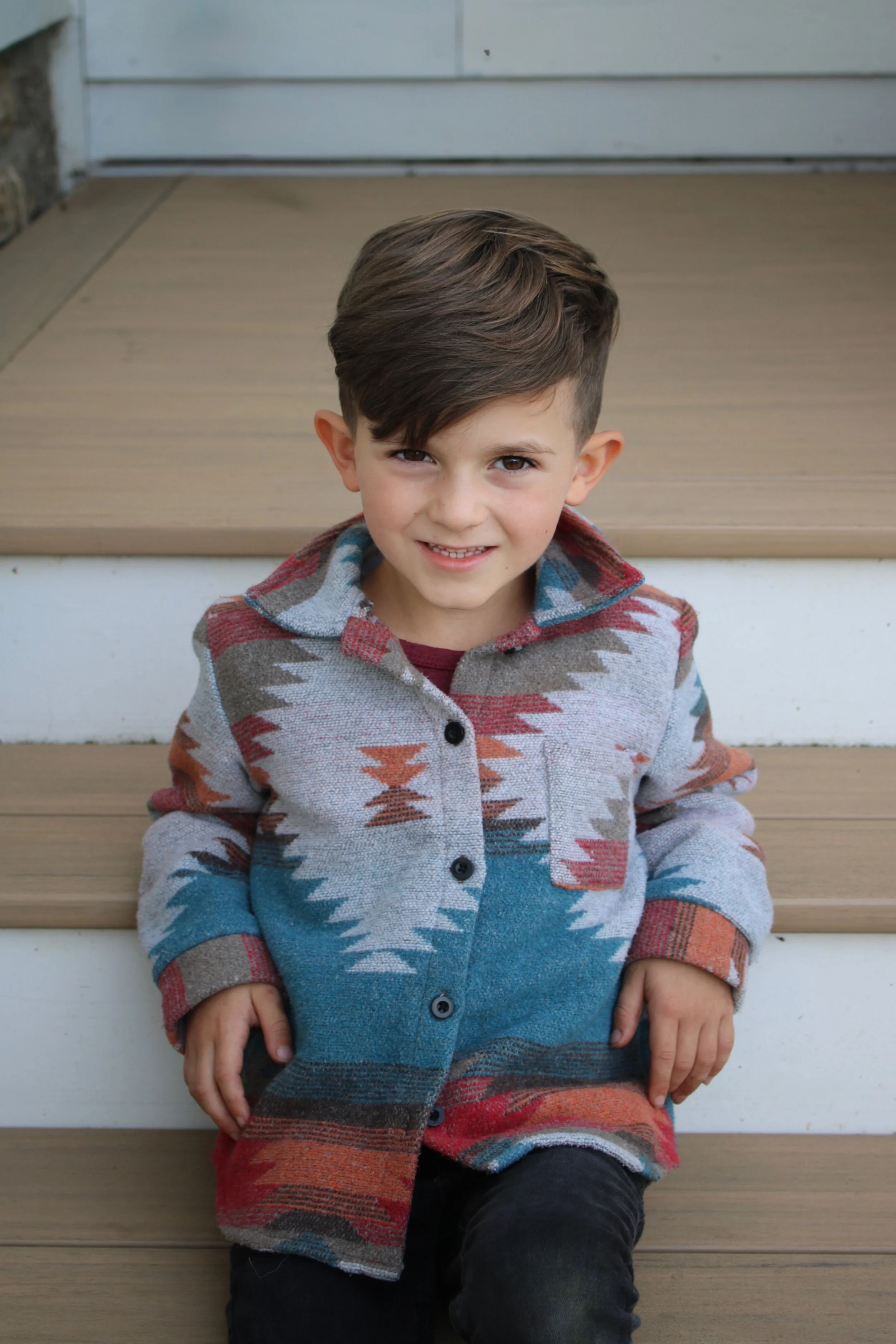 Young boy sitting on outdoor wooden steps, smiling at the camera, wearing a colorful patterned jacket.