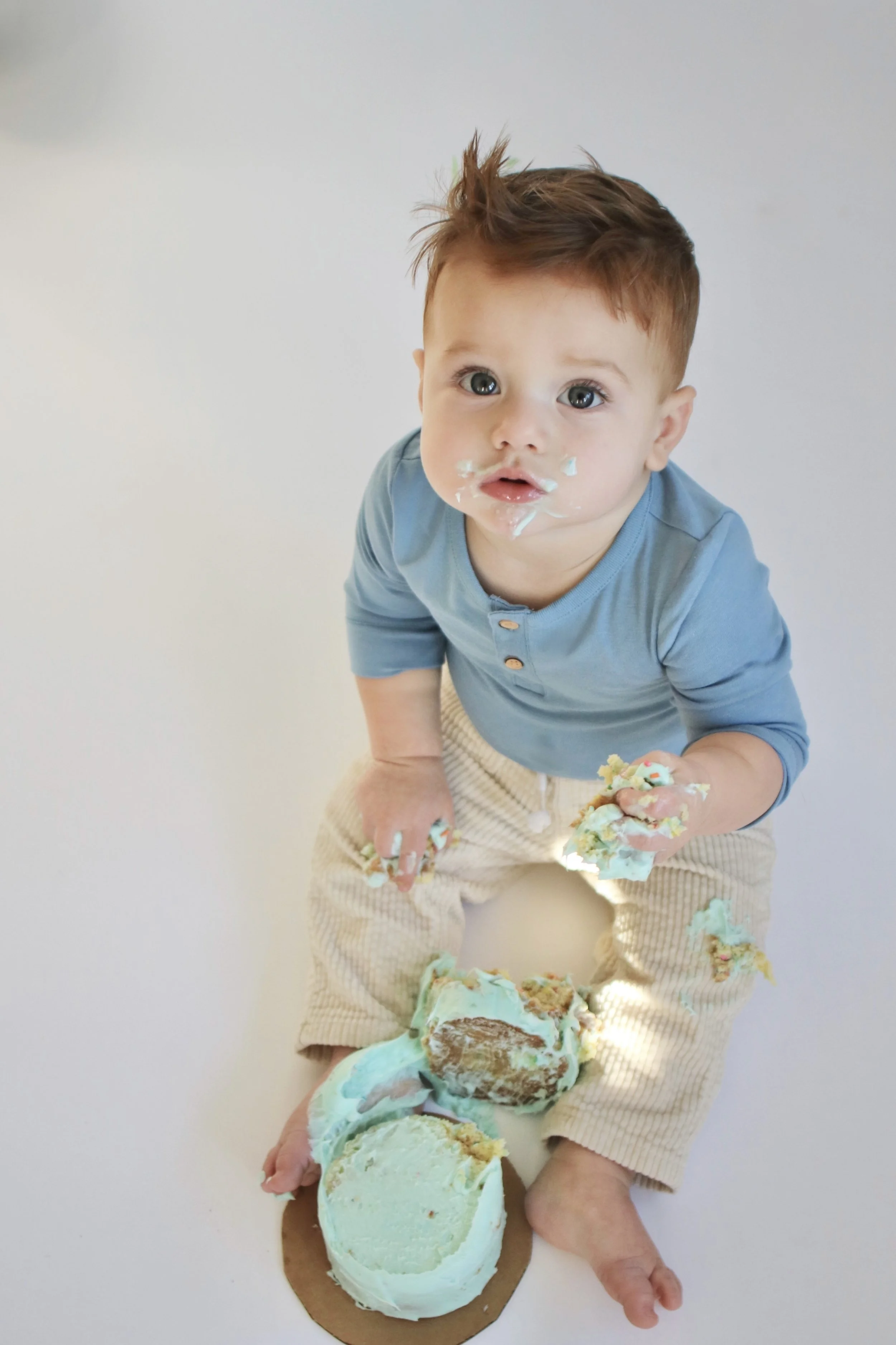 A young toddler with messy face and hands, eating cake with blue and green frosting, sitting on the floor.