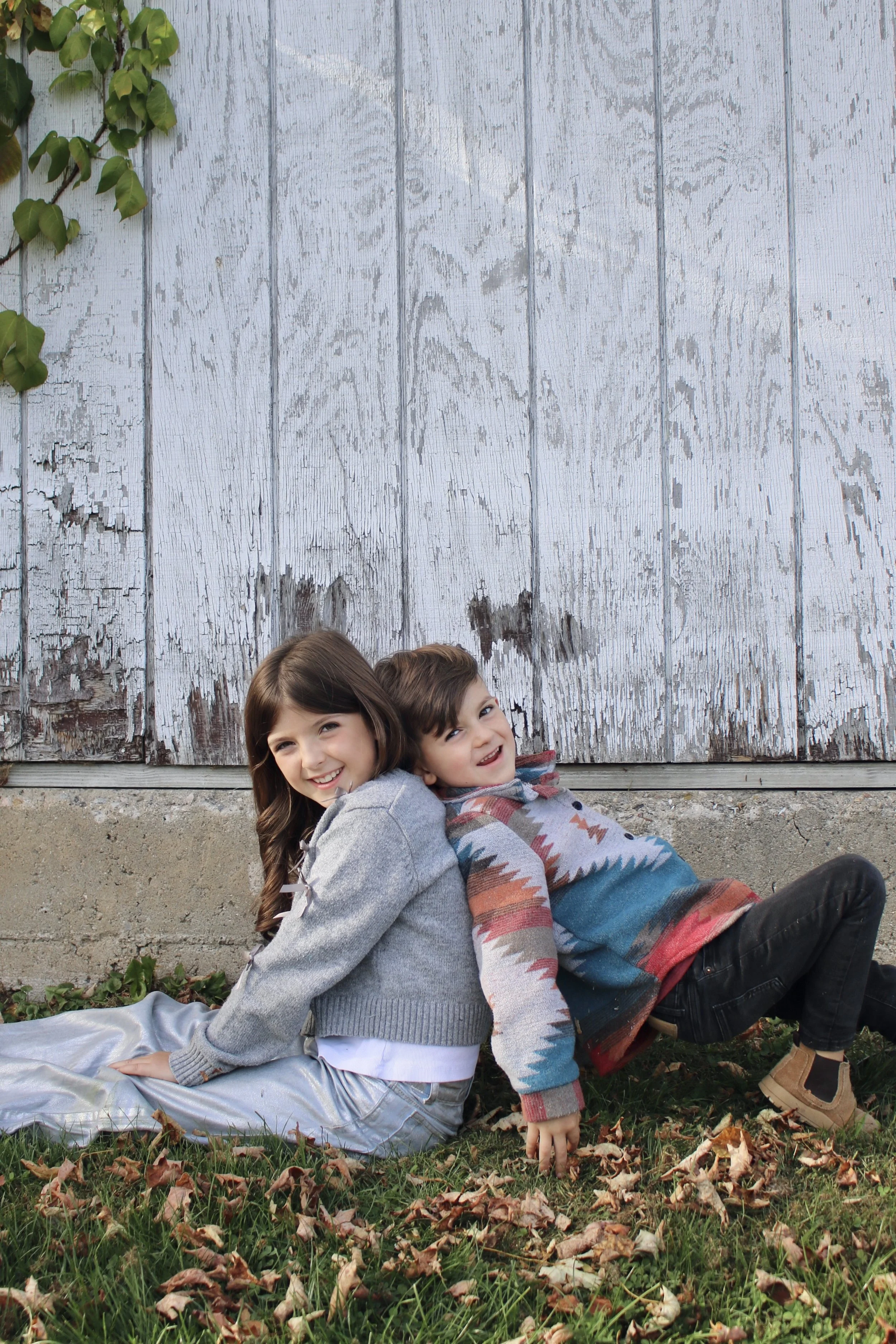Two children, a girl and a boy, sitting back-to-back on the grass with fallen leaves, smiling, outdoors in front of a weathered, white, wooden wall with some climbing plants.