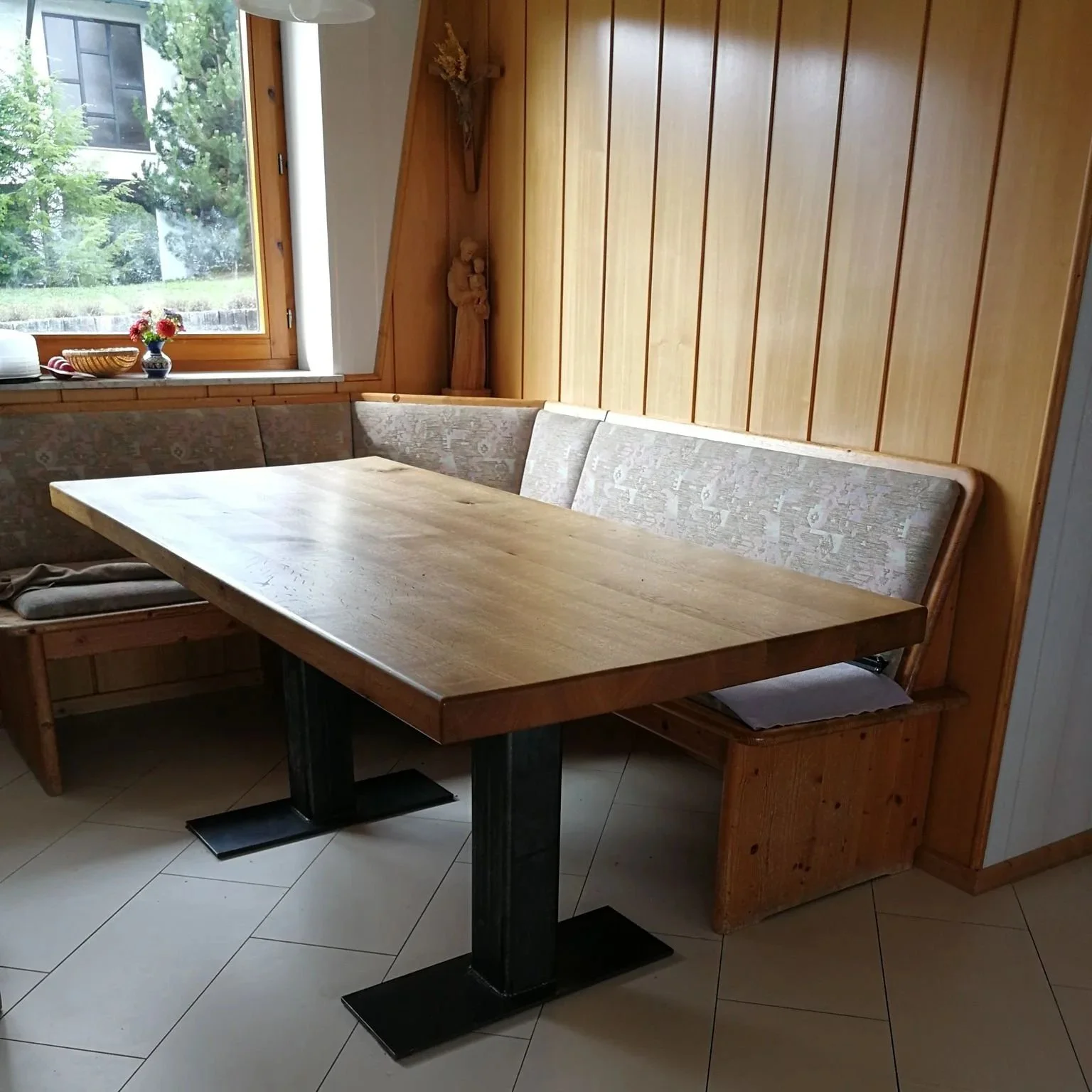 Gemütlicher Holztisch mit angepasster Sitzbank in einer holzverkleideten Ecke, Fenster mit Blick auf grüne Bäume, kleine Dekorationen auf dem Fensterbrett.