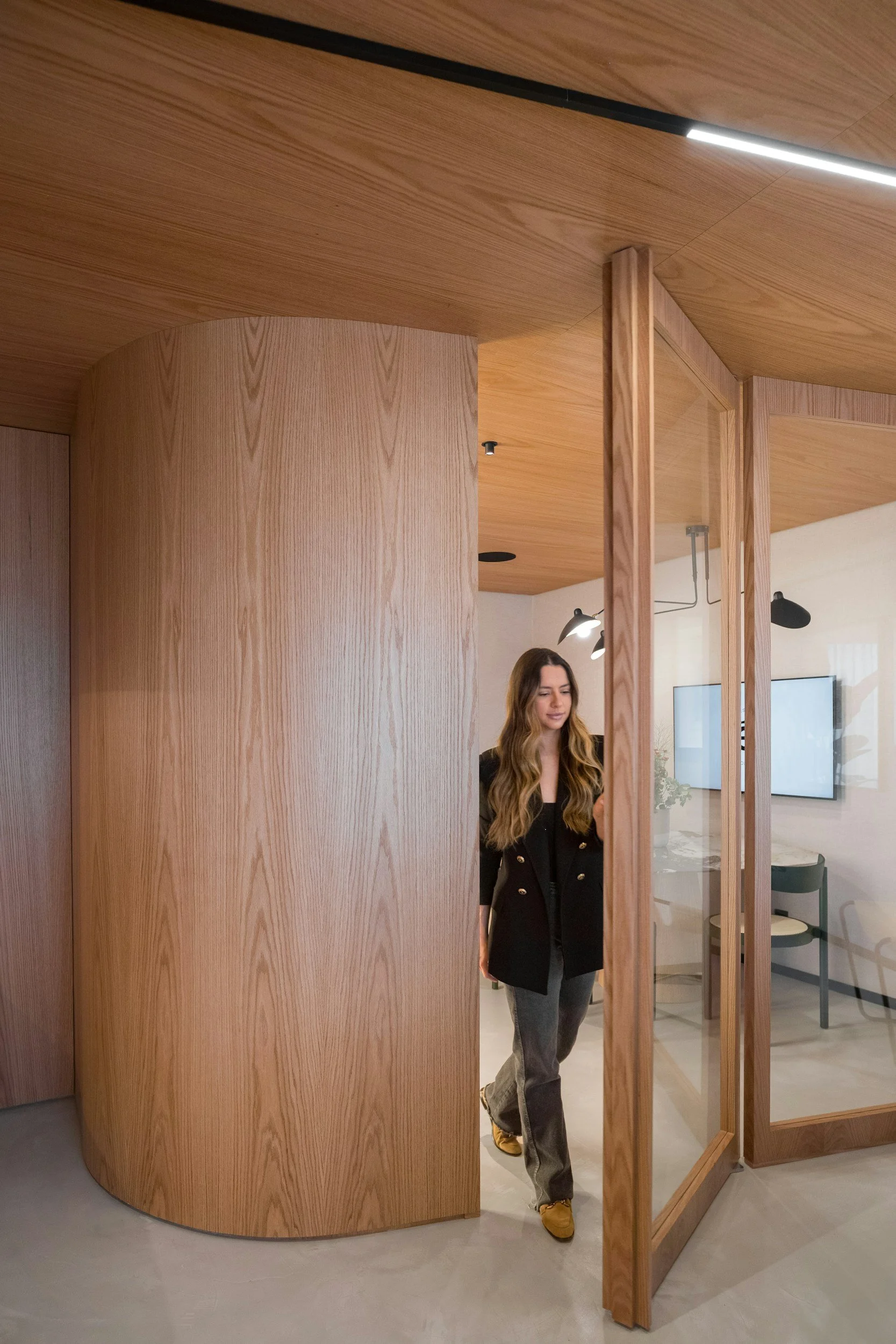 Eine Frau mit langen, gewellten Haaren, trägt eine schwarze Jacke und graue Hosen, geht durch eine moderne, holzverkleidete Türöffnung in einem Büro