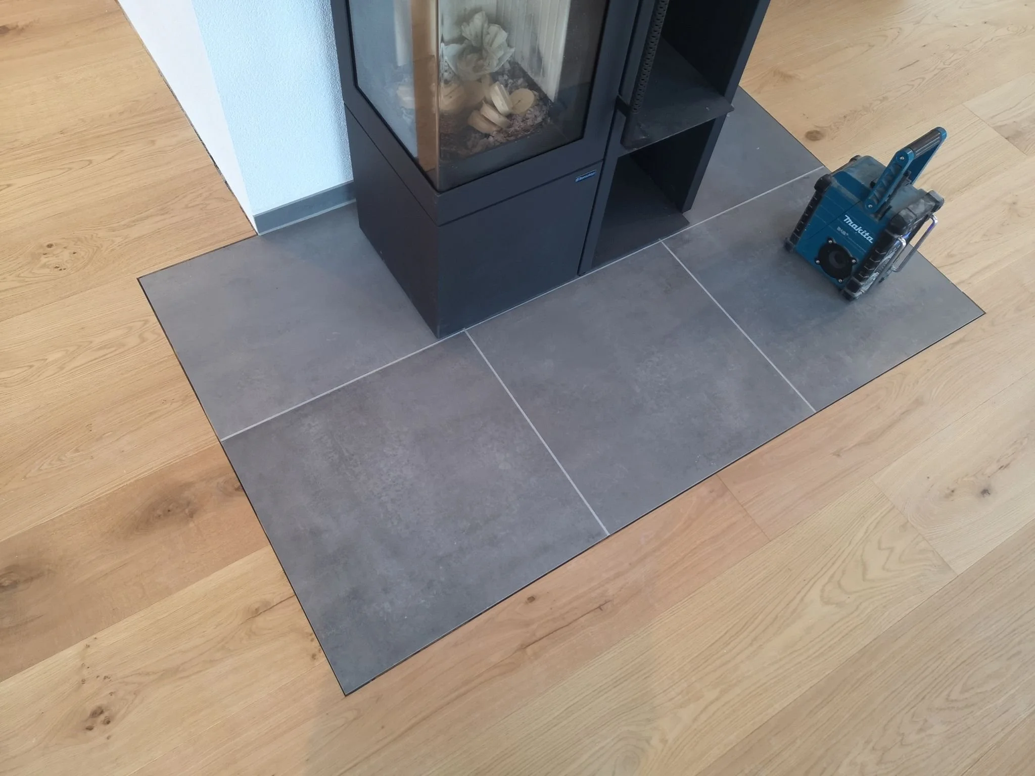 Ein offener Kamin mit schwarzen Metallrahmen auf einer Bodenfliese, daneben ein blauer Werkzeugkoffer.