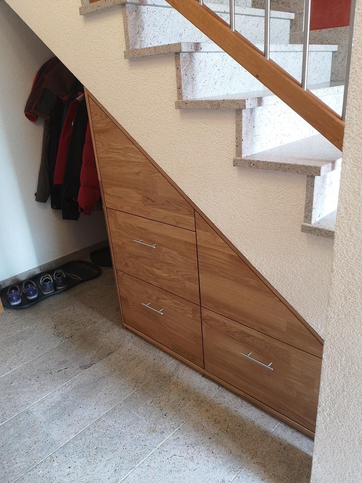 Eingebaute Holzkommode unter der Treppe neben Haken mit Jacken und Schuhen im Flur.