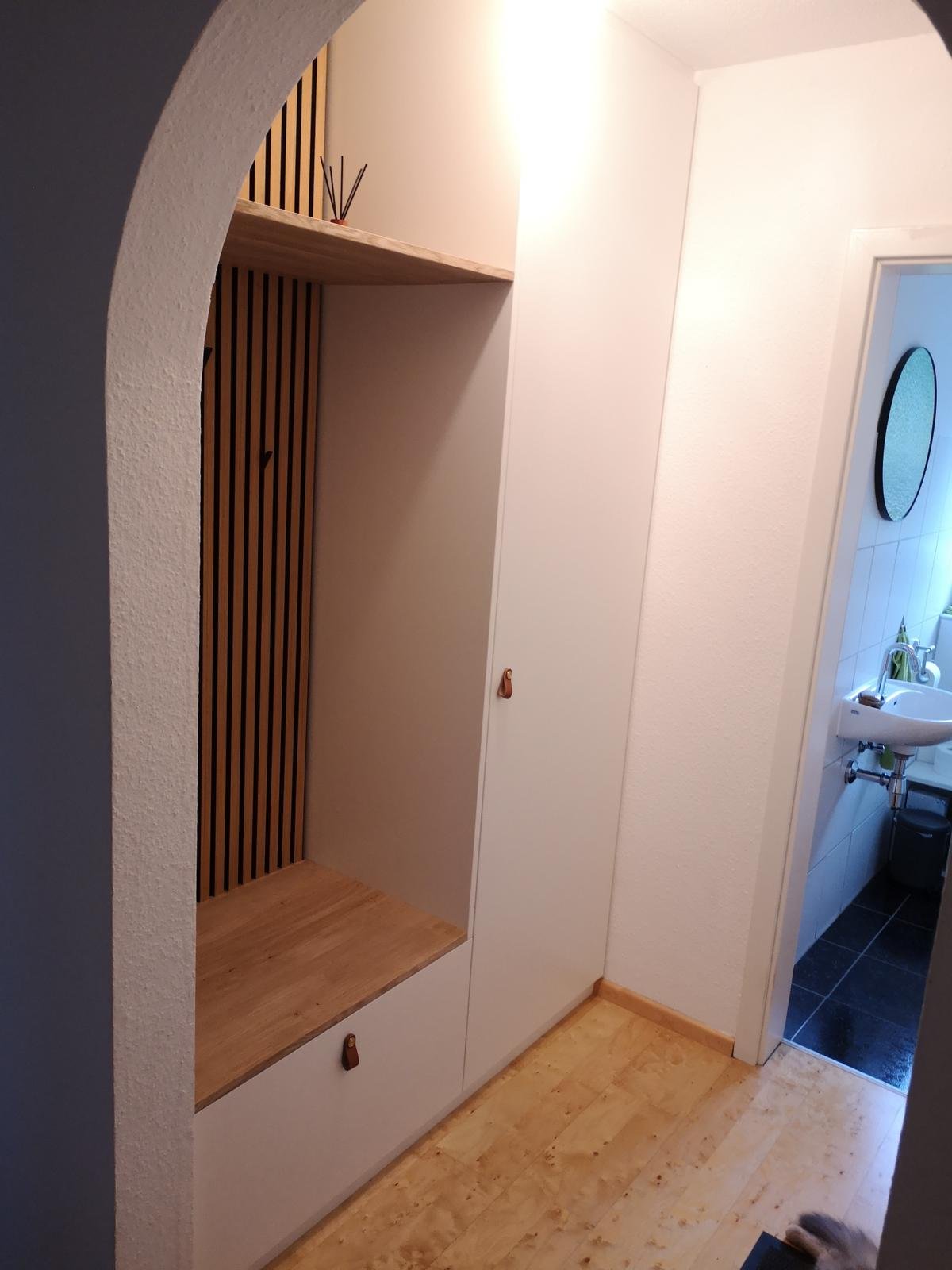 Ein Flur mit einem weißen Schrank, einer Holzregal und einem Holzboden, daneben ein Badezimmer mit Waschbecken und Spiegel.