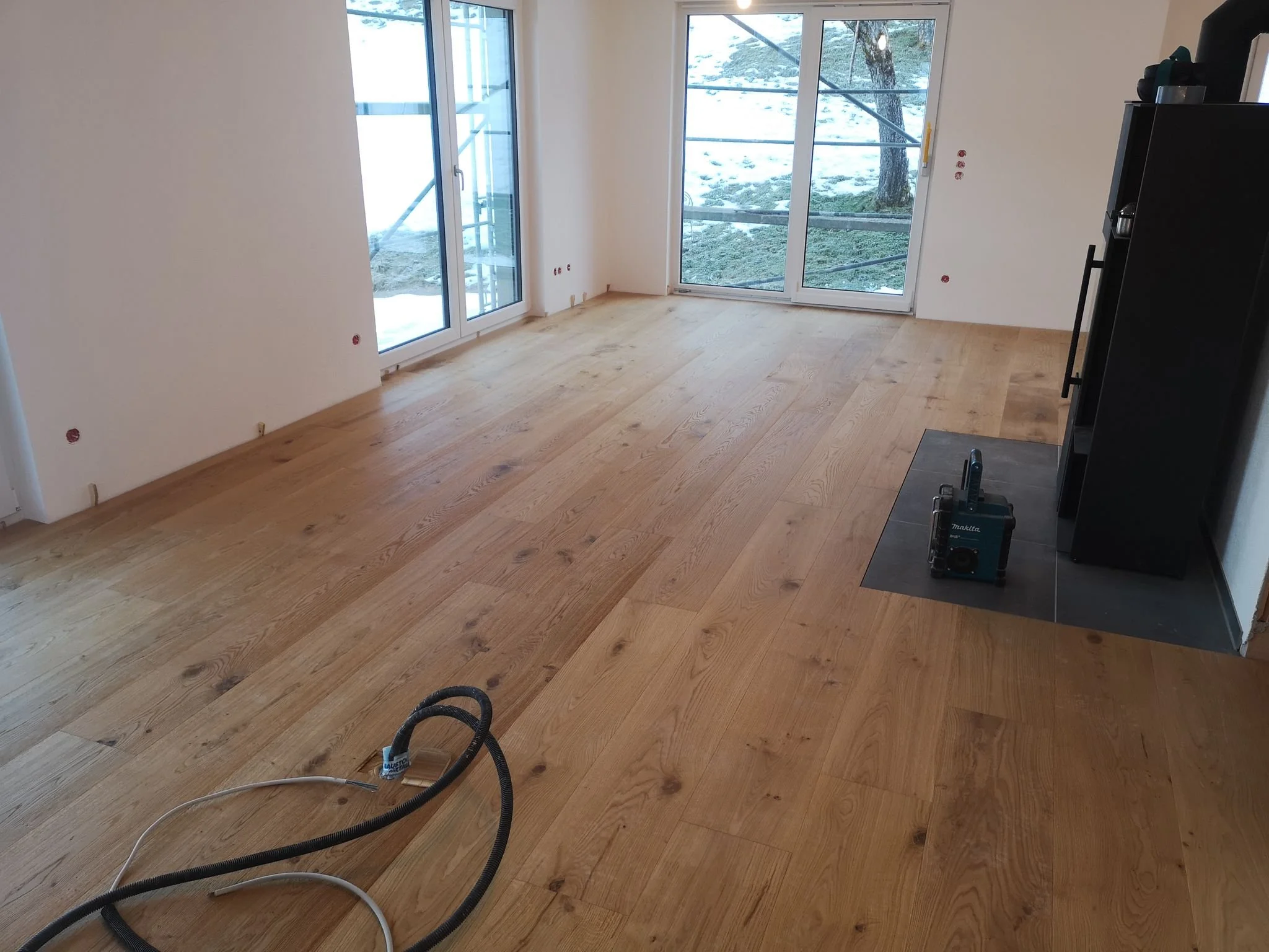 Innenraum eines noch unfertigen Wohnzimmers mit gerade verlegtem Holzparkett, große Glasfenster, die auf eine Schneelandschaft im Freien blicken, und einige Werkzeuge auf dem Boden.