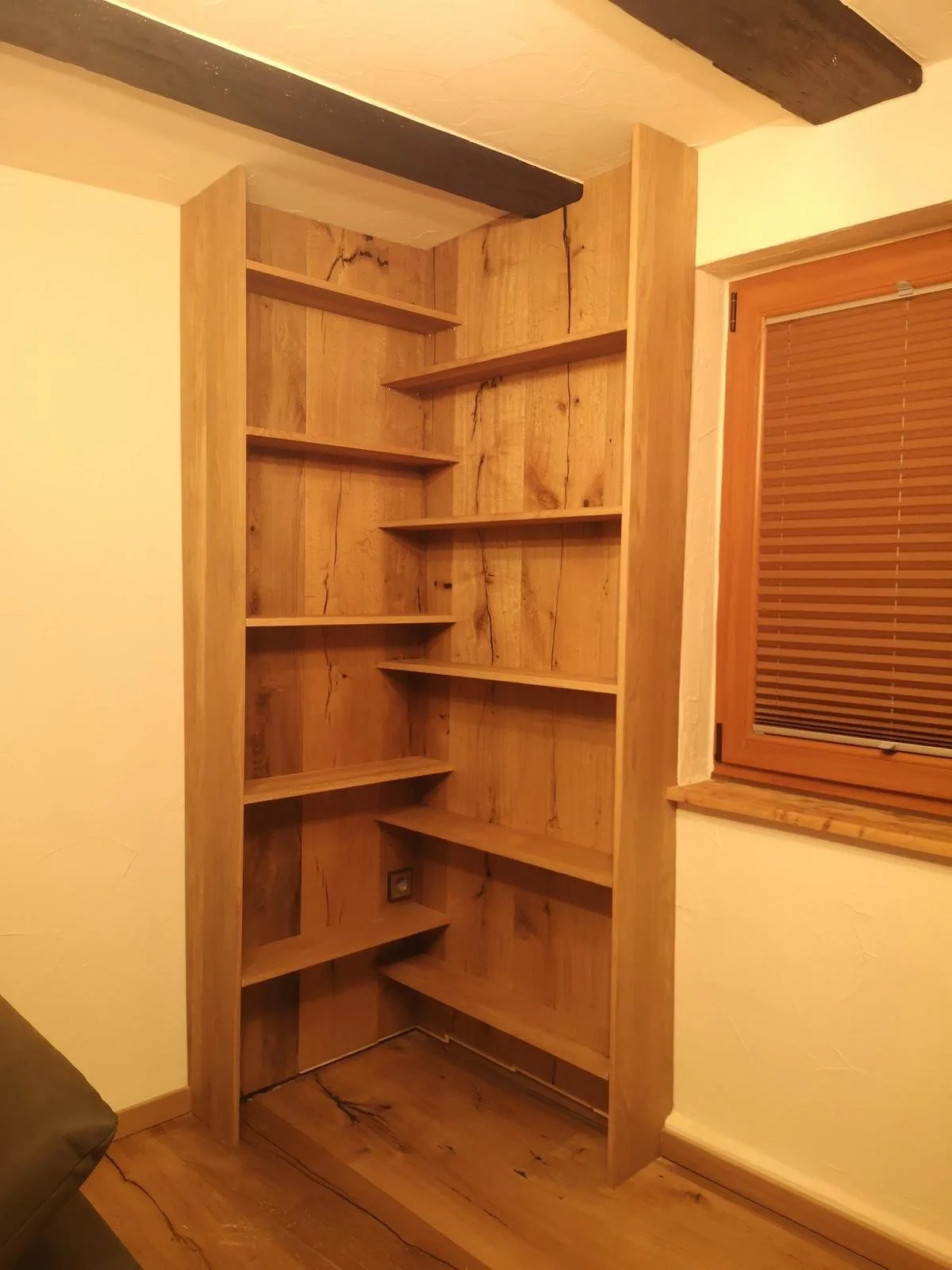 Ein leerer Holzregal mit mehreren Holzregalbrettern, installiert in einer Ecke eines Zimmers mit Holzfußboden, neben einem Fenster mit horizontalen Jalousien.